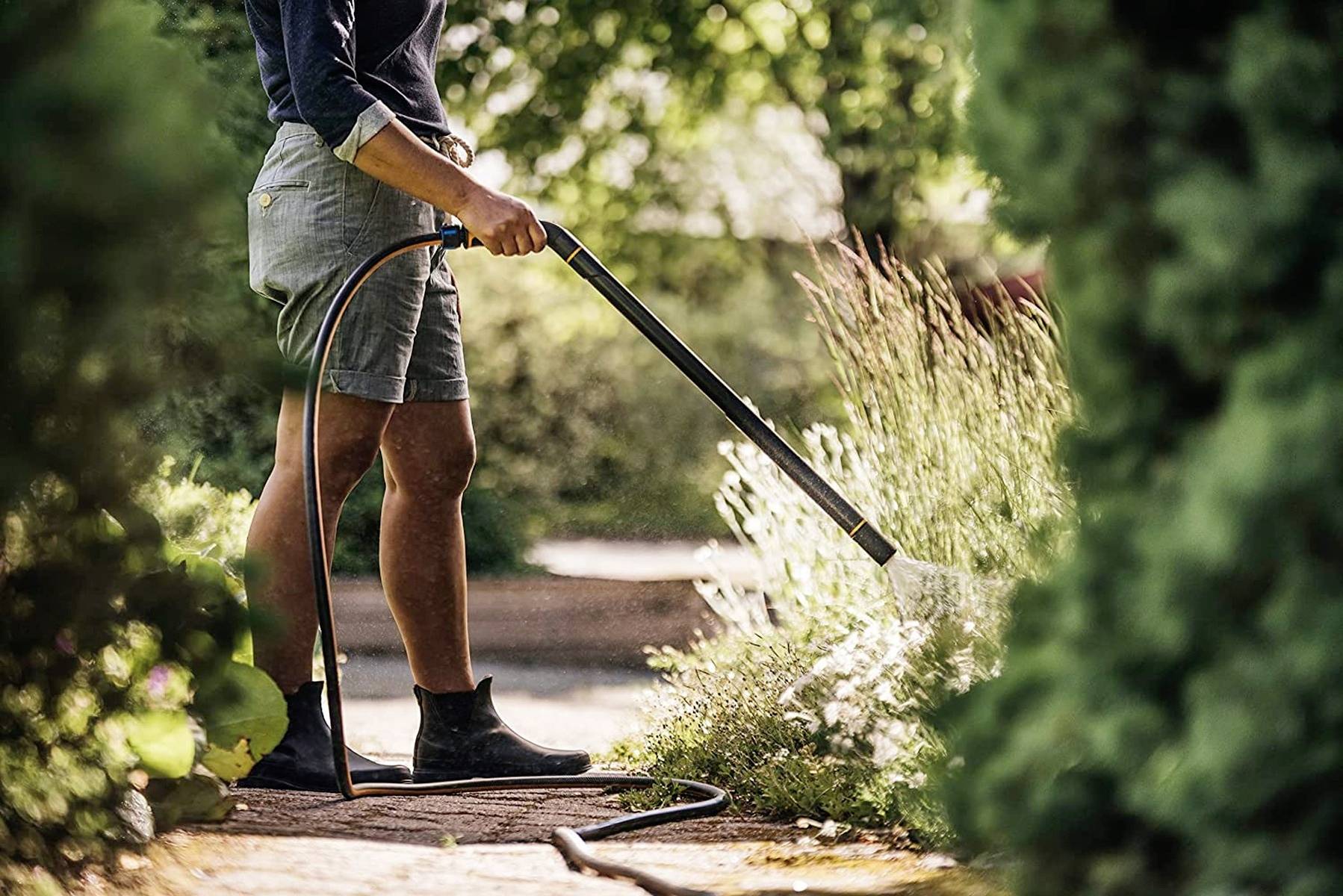 Eine Person gießt Pflanzen mit einem Wasserschlauch in einem grünen Garten. Im Hintergrund sind Bäume und Büsche.