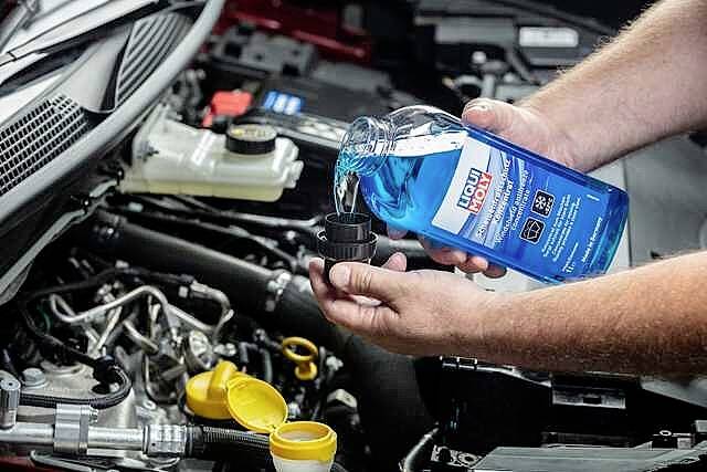 Eine Person füllt blaue Scheibenwischflüssigkeit aus einer Flasche in den Behälter im Motorraum eines Autos.