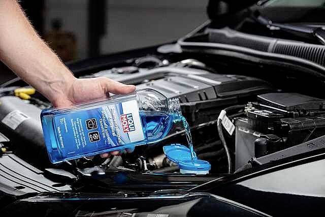 Eine Person füllt blaue Scheibenwaschflüssigkeit in den Behälter eines offenen Autotanks. Der Motorraum ist sichtbar.