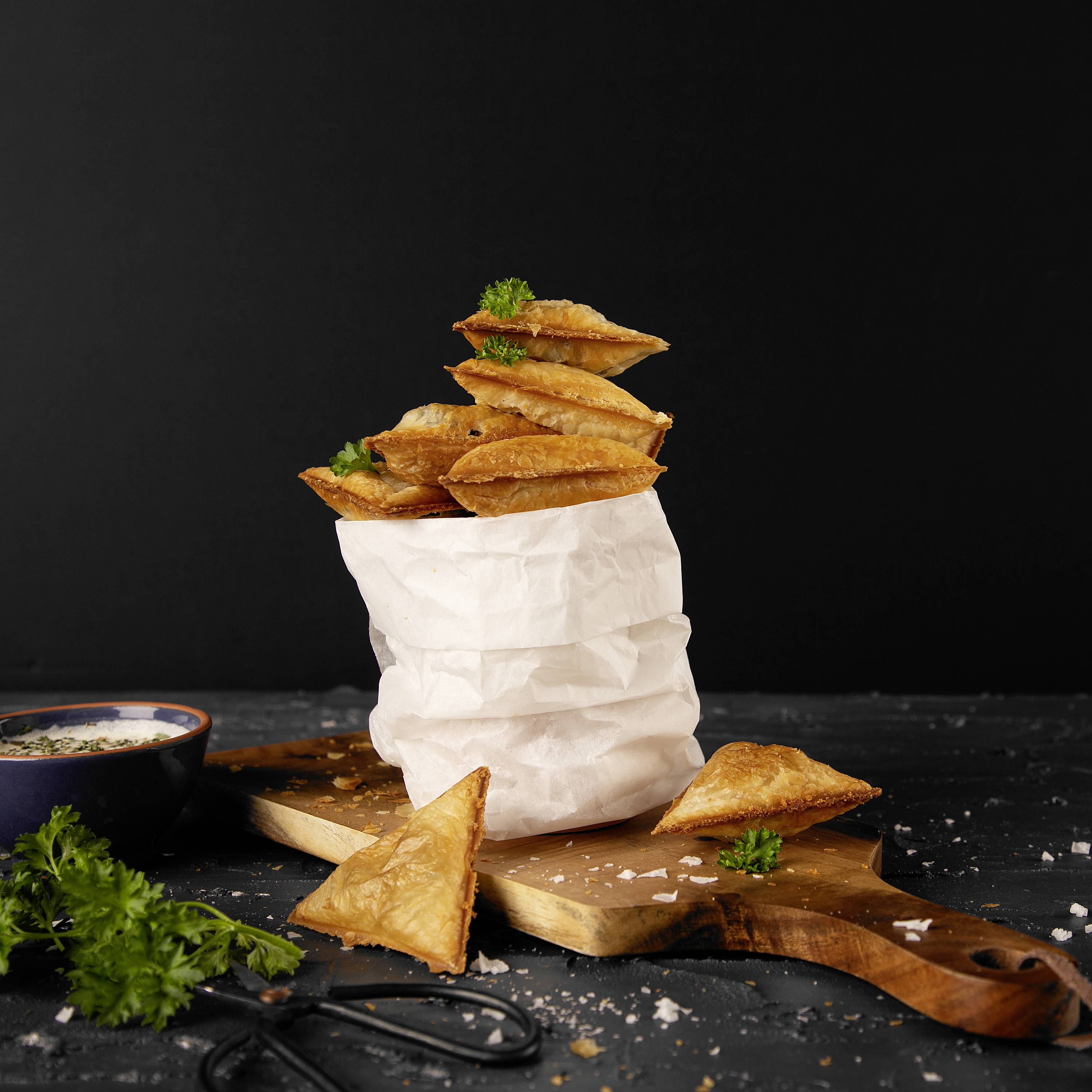 Ein Stapel Plundergebäck auf braunem Papier, dekoriert mit Kräutern, liegt auf einem Schneidebrett. Eine Schüssel mit Soße und frische Kräuter sind daneben platziert.