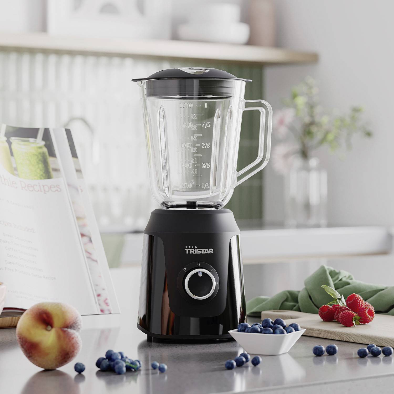 Ein schwarzer Mixer mit Messbecher steht auf einem Küchenarbeitsplatz. Daneben liegen Heidelbeeren, Erdbeeren und ein Pfirsich. Im Hintergrund ist ein offenes Kochbuch sichtbar.