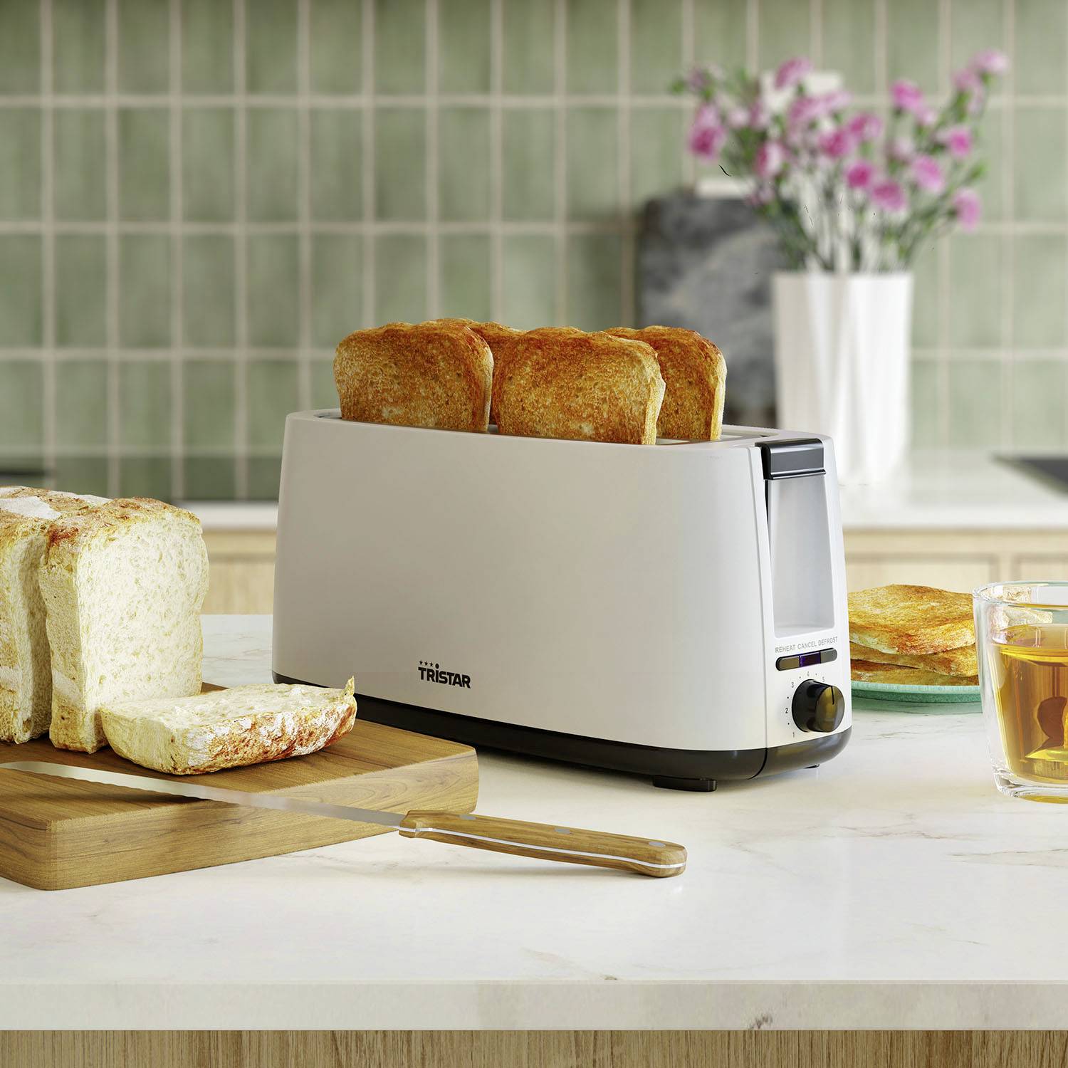 Ein Toaster mit zwei gebräunten Toasts steht auf einer Küchenarbeitsplatte. Daneben liegen geschnittenes Brot und ein Glas Tee.