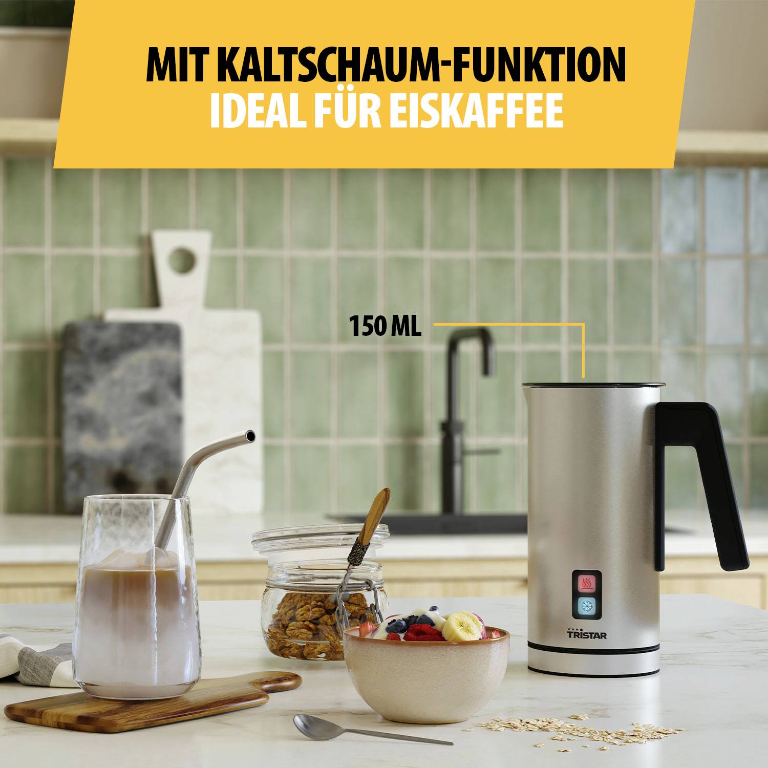 Elektrischer Milchaufschäumer auf Küchentisch, daneben Glas Eiskaffee mit Strohhalm, Müslischale und Käsestücke im Hintergrund.