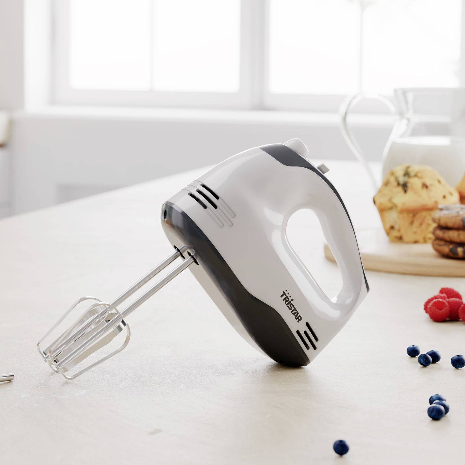 Ein Handmixer auf einer Küchenarbeitsplatte neben einem Glas Milch und Keksen. Durch das Fenster fällt Tageslicht herein.