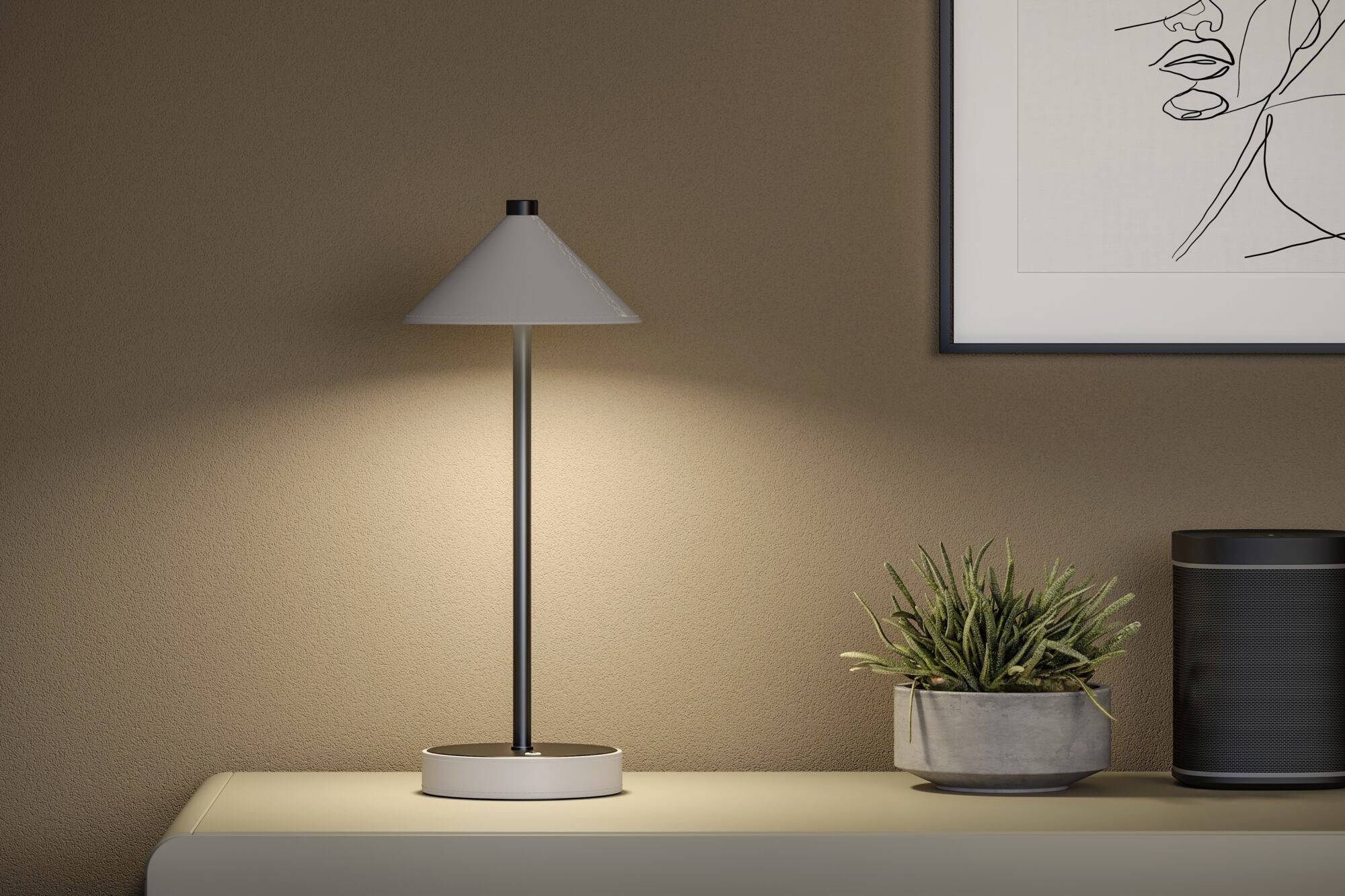 Eine Tischlampe leuchtet sanft auf einem Tisch, neben einem kleinen Pflanztopf. An der Wand hängt ein lineares Kunstwerk.