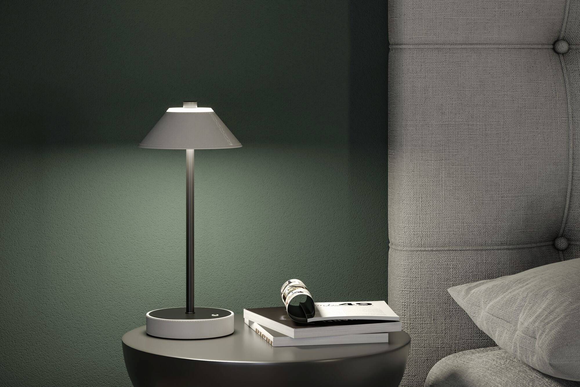 Eine Nachttischlampe leuchtet auf einem runden Tisch neben einem Bett. Auf dem Tisch liegen Bücher und eine gerollte Zeitschrift.