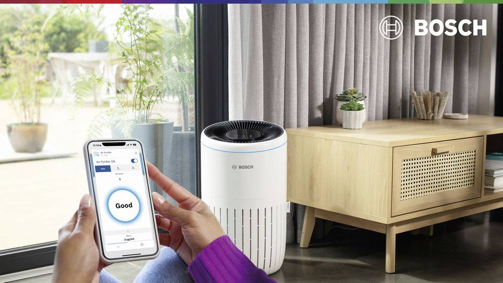 Eine Person überprüft die Luftqualität auf einem Smartphone. Ein Luftreiniger der Marke Bosch steht daneben im modernen Wohnzimmer.