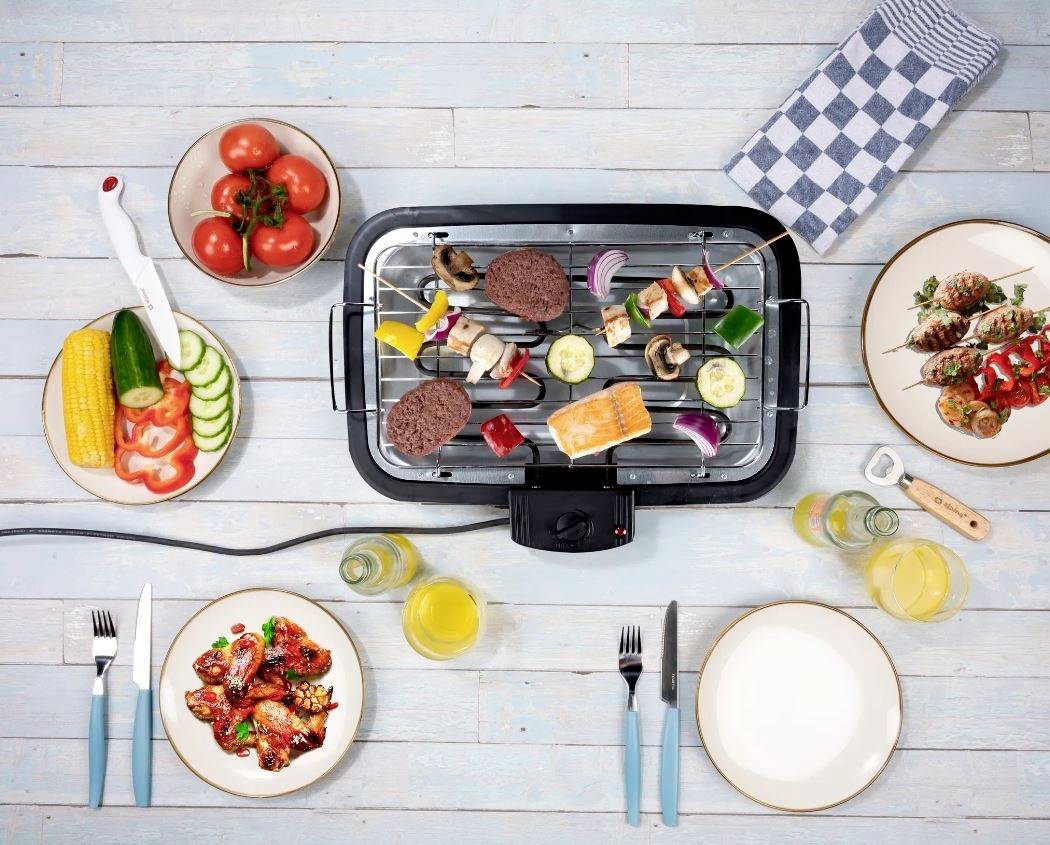 Ein Tischgrill auf einem Holztisch mit Gemüse, Fleisch und Spießen. Umgeben von Tellern, Tomaten, Gläsern mit Getränken und Besteck.