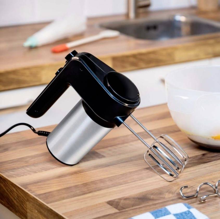 Küchenmaschine auf hölzerner Arbeitsfläche, mit Schneebesenaufsätzen. Nebenbei eine weiße Schüssel, bereit zum Backen oder Kochen.