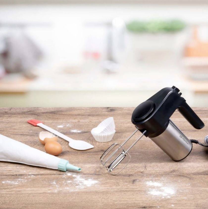 Ein Handmixer liegt auf einem Holztisch neben einem Spritzbeutel, einem Ei, einem Teiglöffel und einer Papierbackform in einer Küche.