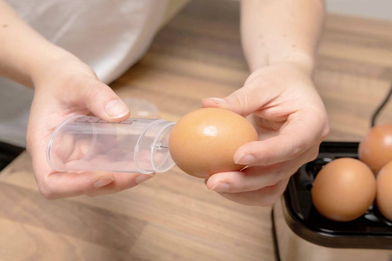 Eine Person hält ein Ei in der Hand und bereitet es vor, in einen Eierkocher zu legen. Ein transparenter Messbecher wird benutzt.