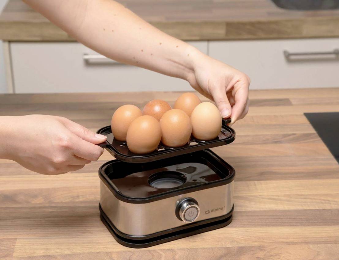 Eine Person legt ein Tablett mit sechs Eiern auf einen Eierkocher in einer Küche. Der Eierkocher steht auf einer Holzarbeitsplatte.