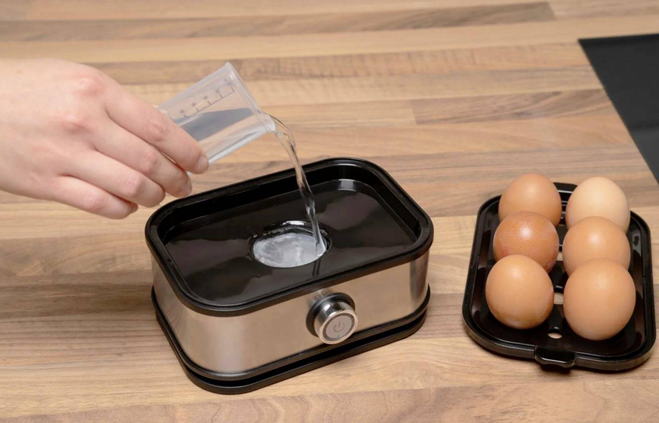 Eine Hand gießt Wasser in einen Eierkocher, daneben liegen sechs braune Eier auf einem Eierhalter auf einer Holzarbeitsplatte.
