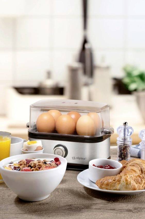 Küche mit einem Eierkocher voller Eier, Müsli-Schale, Croissant, Saft und Gewürzen auf einem Tisch.