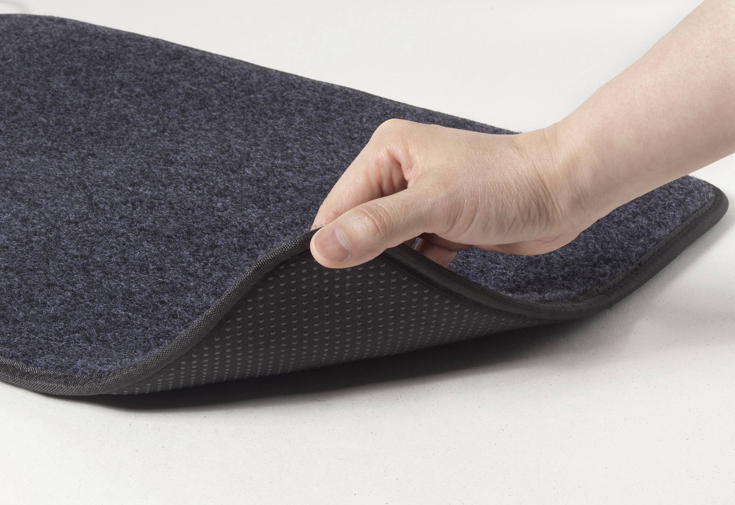 Eine Hand hebt die Ecke einer grauen Türmatte und enthüllt eine rutschfeste Rückseite.
