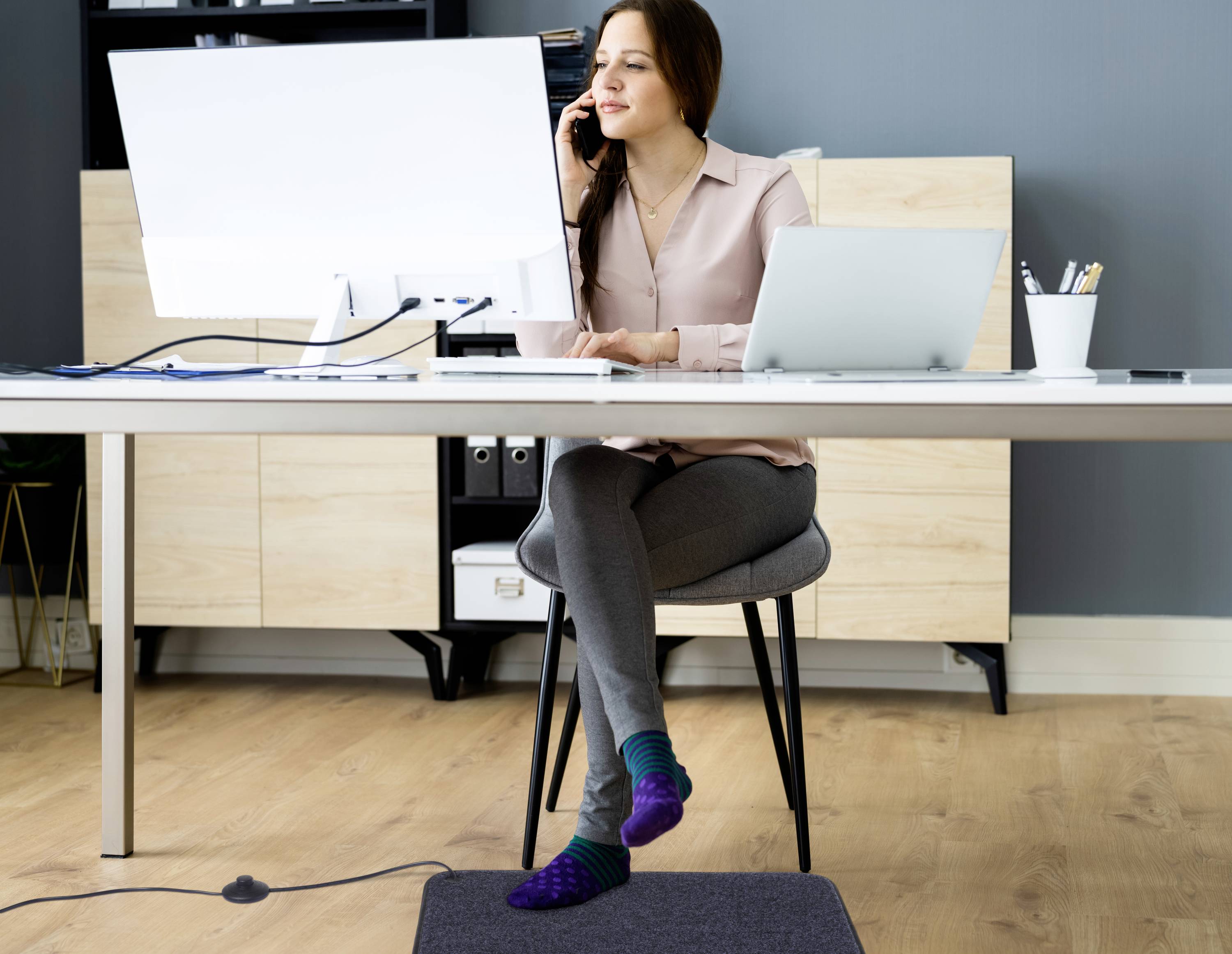 Eine Frau sitzt an einem Schreibtisch mit einem Telefon am Ohr und arbeitet an einem Computer. Sie trägt Socken und hat ein Bein übergeschlagen, wirkt konzentriert.
