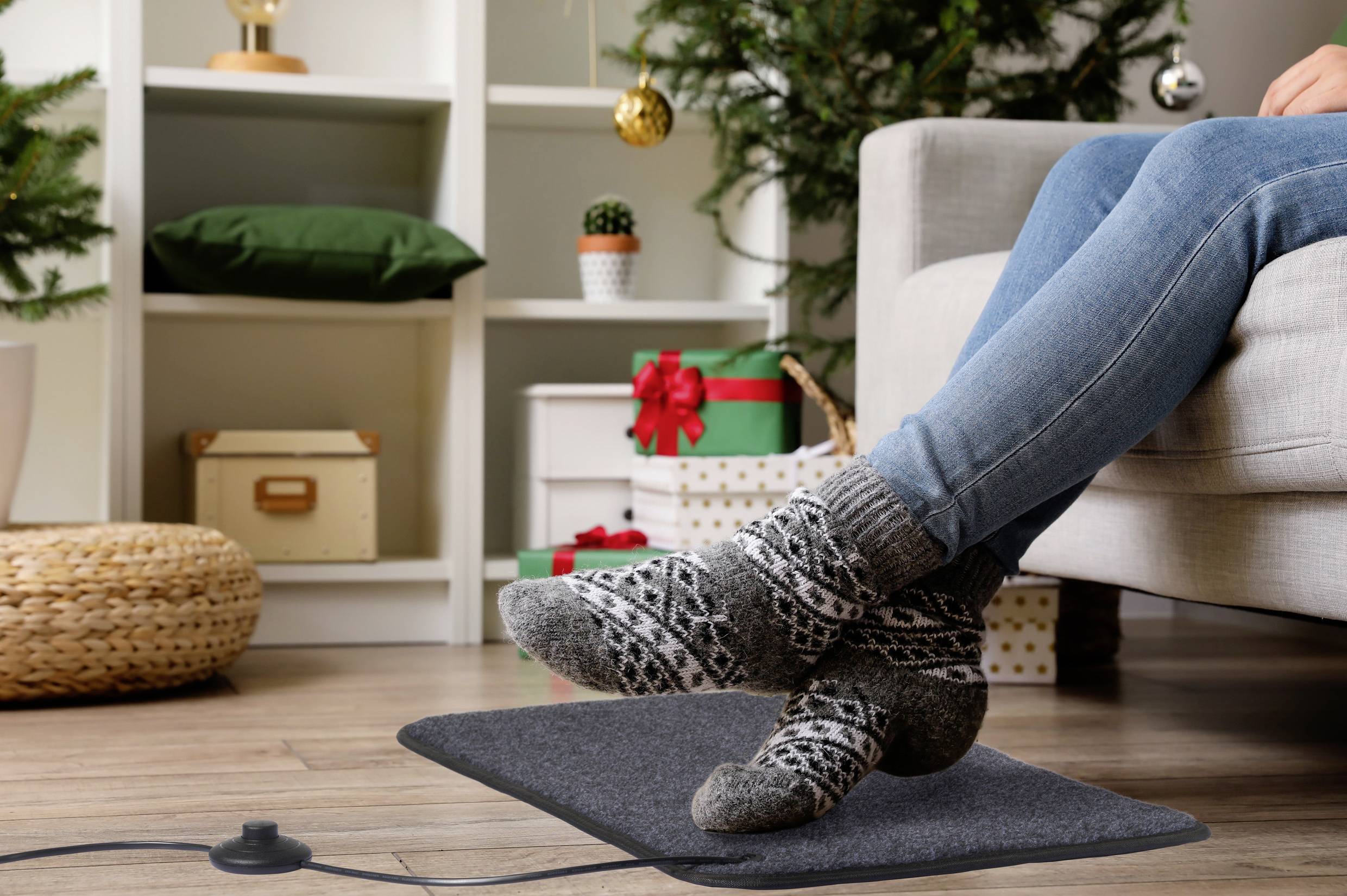 Eine Person mit gemütlichen Socken ruht die Füße auf einer Heizmatte, während Weihnachtsgeschenke und ein geschmückter Baum im Hintergrund eine festliche Atmosphäre schaffen.