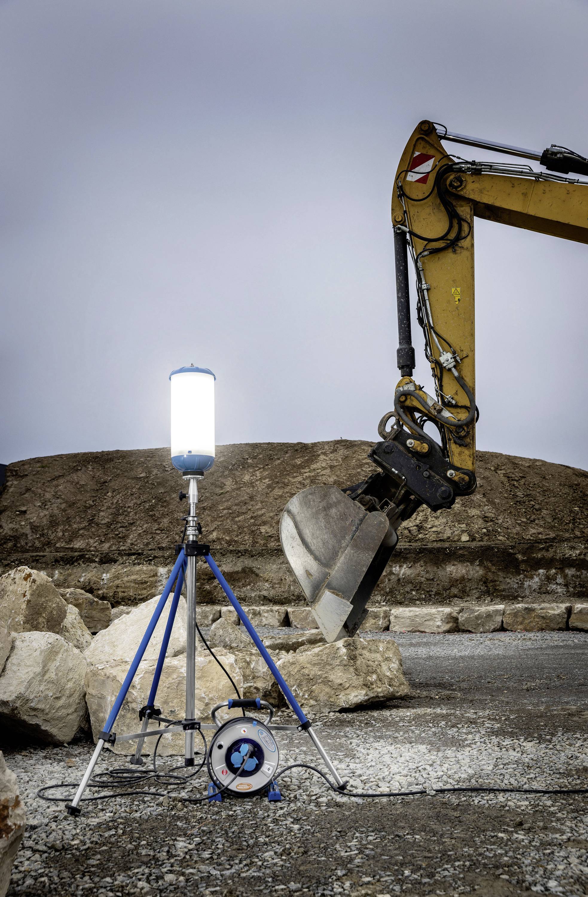 Eine Baustelle zeigt einen beleuchteten Scheinwerfer auf einem Stativ neben einem Baggerarm mit Schaufel, umgeben von Steinen.
