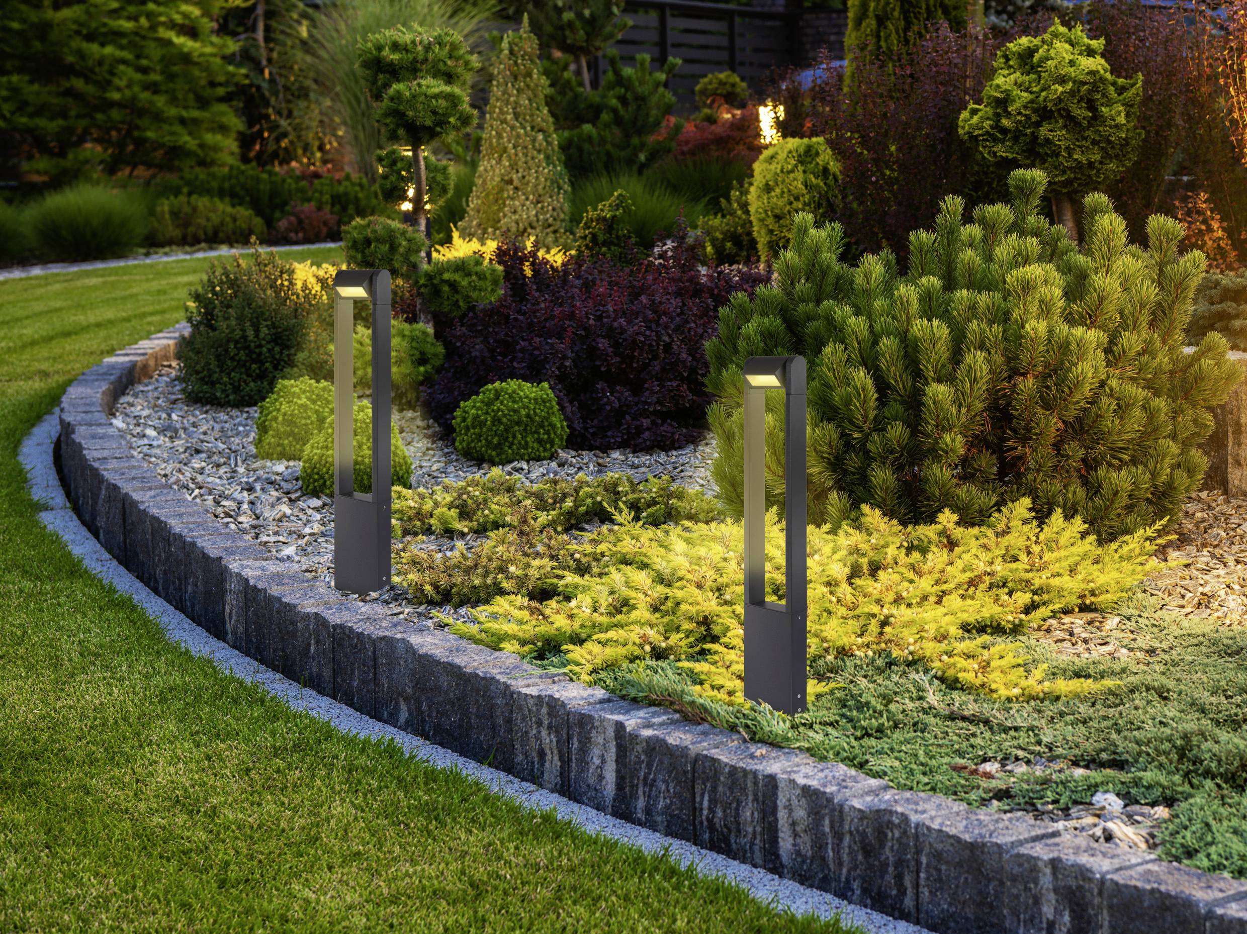 Ein landschaftlich gestalteter Garten mit sorgfältig geschnittenen Sträuchern und dekorativen Beleuchtungskörpern entlang einer Steinbegrenzung, umgeben von grünem Rasen.