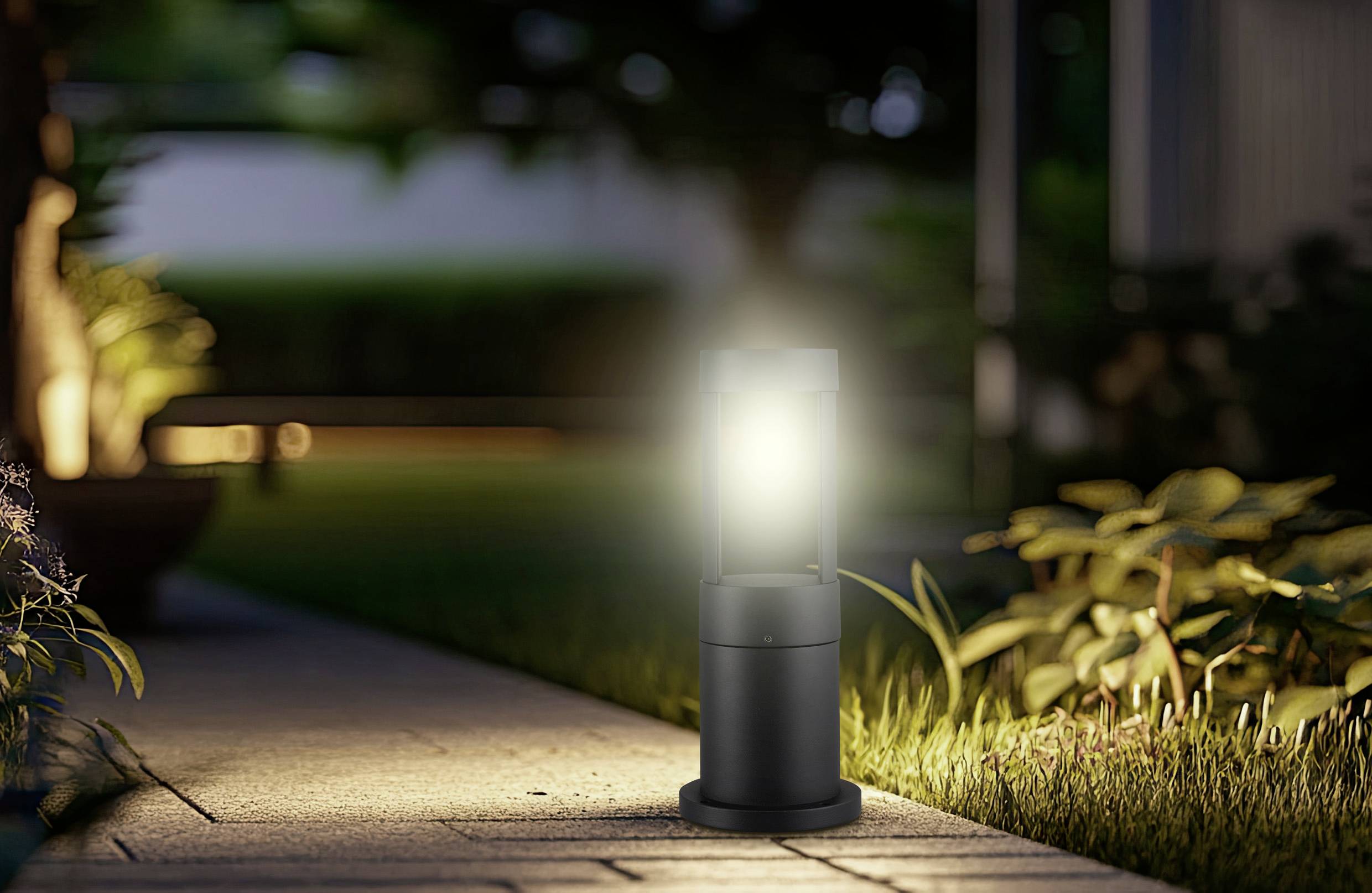 Beleuchteter Wegbeleuchtungslicht in einem Garten am Abend, umgeben von Gras und Pflanzen, der eine warme und einladende Atmosphäre schafft.