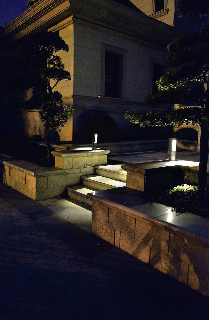 Nächtliche Außenszene mit sanft beleuchteten Steinstufen, die zu einem Gebäudeeingang hinaufführen, umgeben von kleinen Bäumen und Büschen.