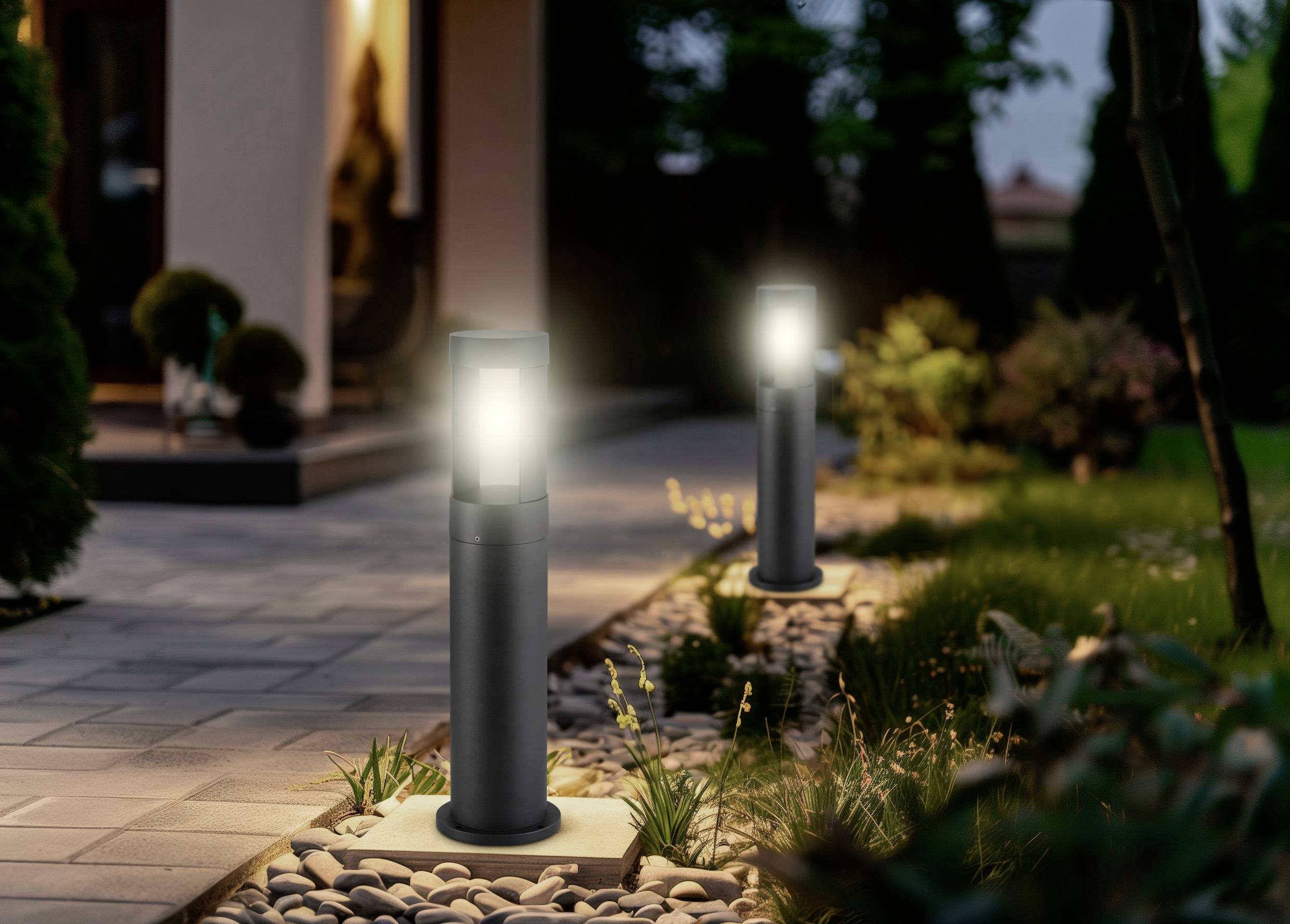 Zwei moderne Außenleuchten beleuchten einen mit Steinen und Gras gesäumten Weg, der zu einem Haus mit warmem Lichtschein im Inneren führt.