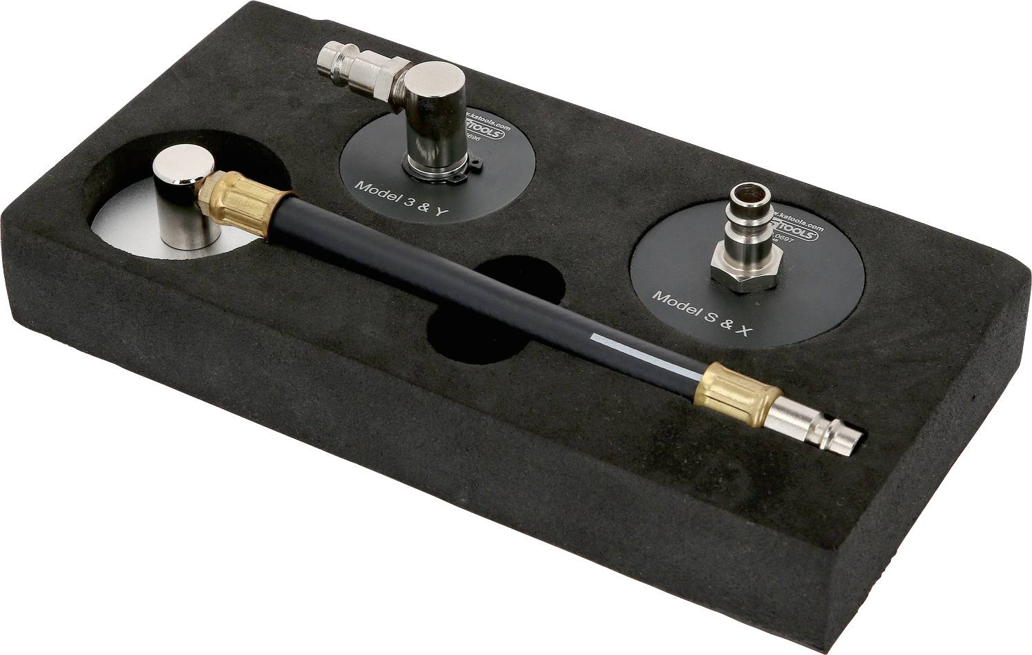 Eine schwarze Schaumstoffverpackung enthält zwei metallische Bauteile, die mit 'Model 3 & Y' und 'Model S & X' beschriftet sind, sowie einen schwarzen Schlauch mit goldenen Anschlüssen.