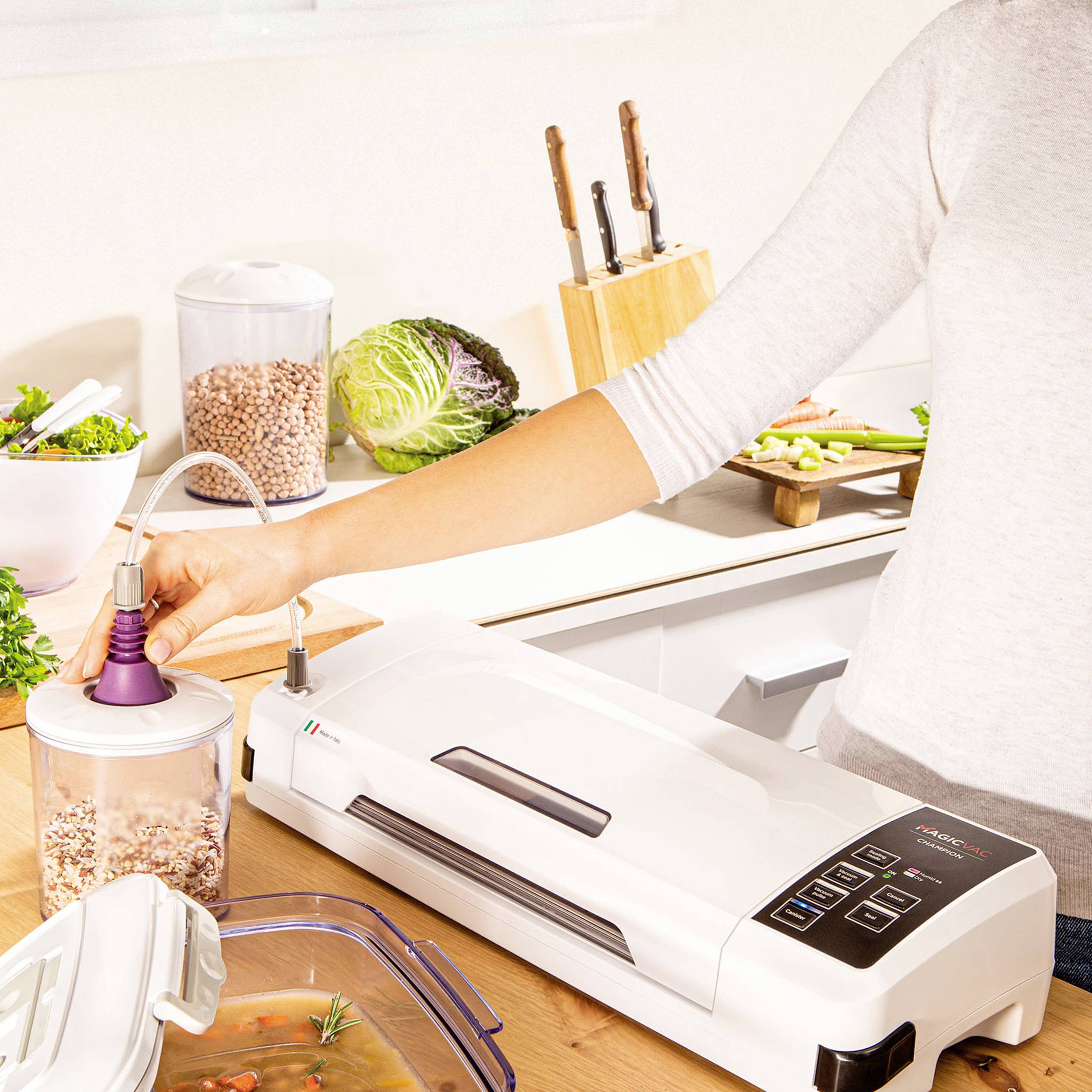 Eine Person benutzt ein Vakuumiergerät, um Lebensmittel in einem Behälter zu versiegeln. Im Hintergrund liegen frisches Gemüse und ein Schneidebrett auf der Küchentheke.