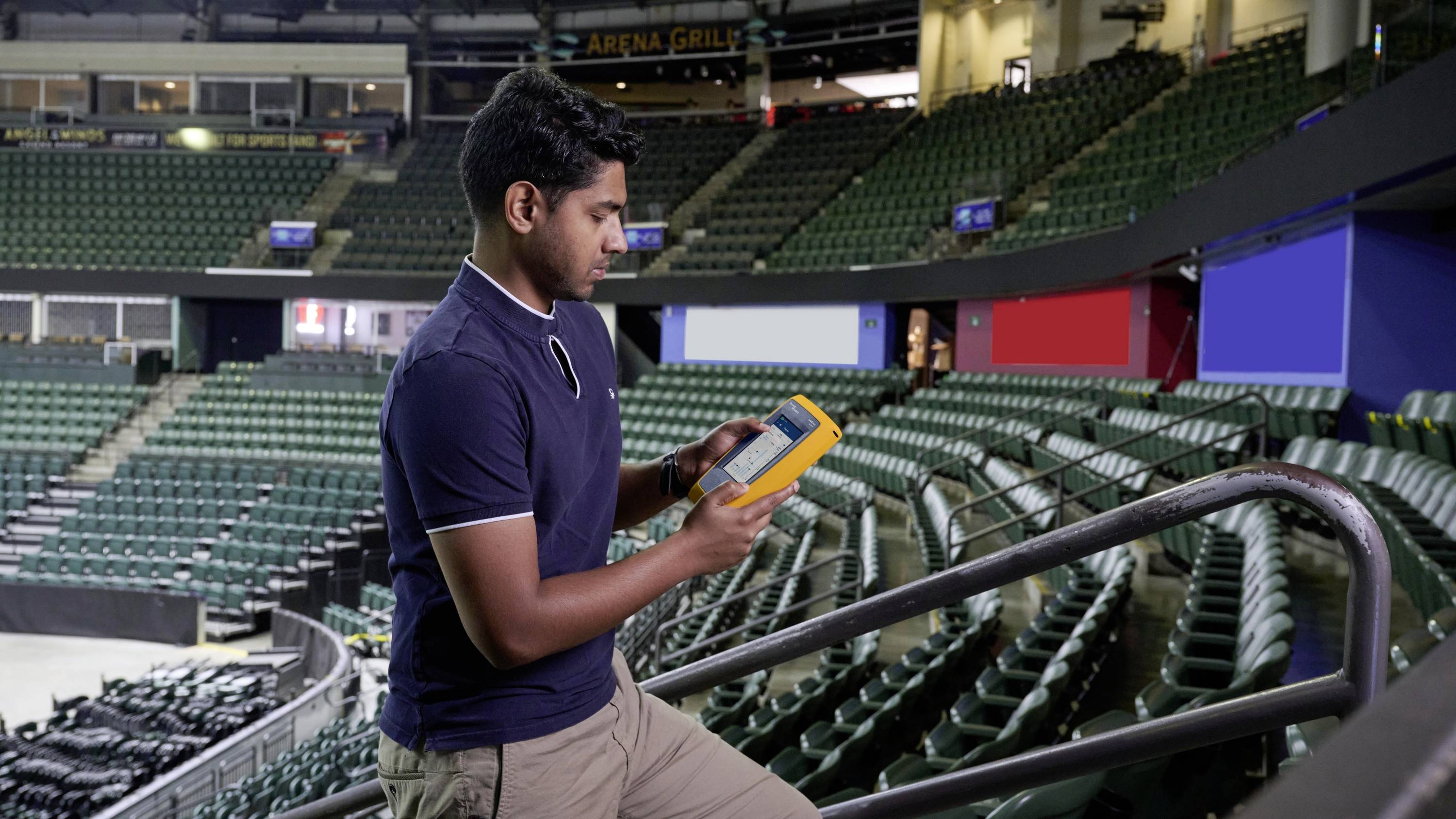 Eine Person steht in einer leeren Arena und betrachtet ein technisches Gerät. Die Sitzreihen im Hintergrund sind leer.