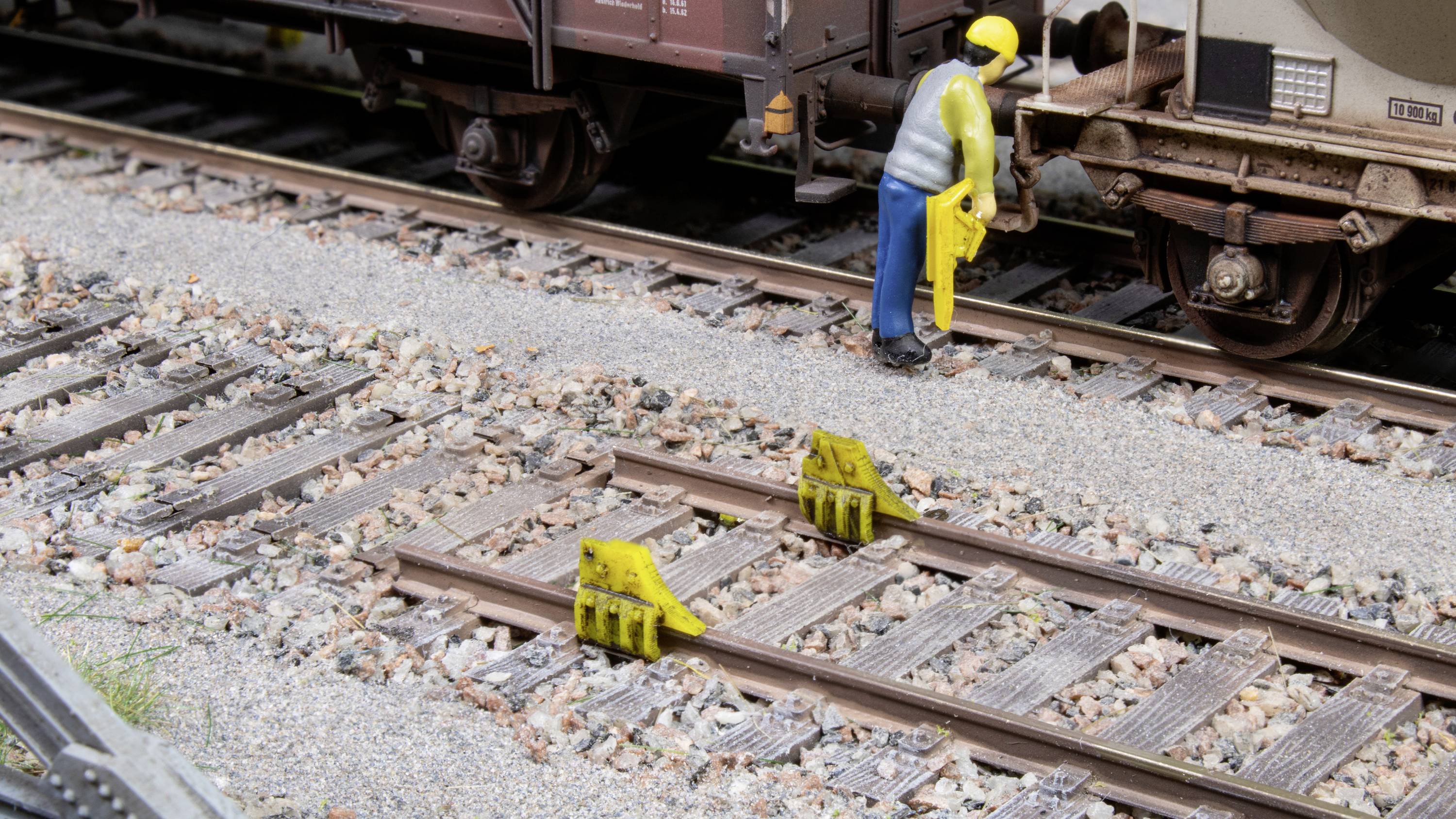 Ein Miniatureisenbahnarbeiter steht zwischen Schienen und hält ein gelbes Werkzeug. Schienenbegrenzungen sind auf den Gleisen sichtbar.