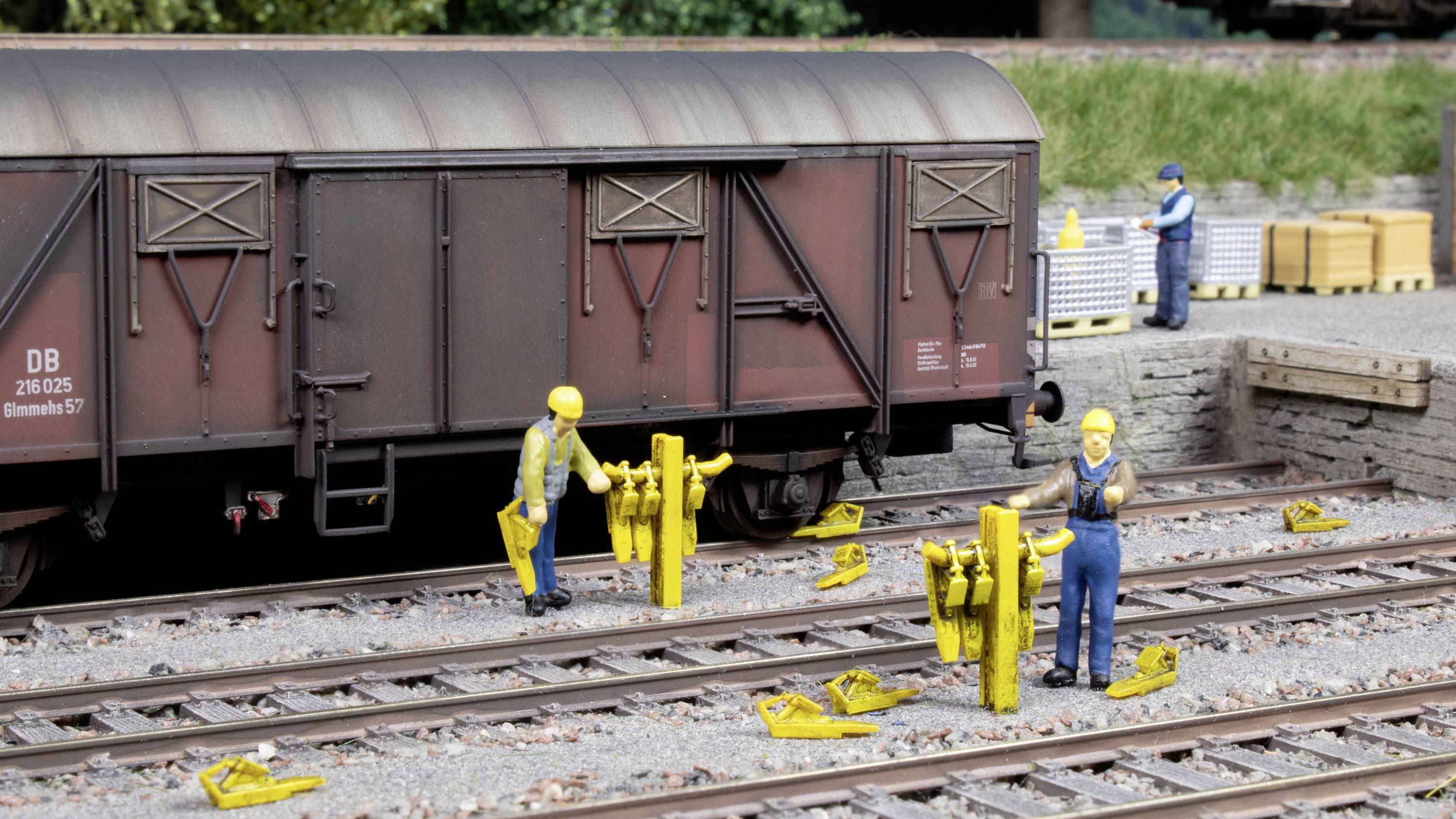 Eine Modelleisenbahn-Szene zeigt Arbeiter, die mit gelben Werkzeugen an Eisenbahnschienen arbeiten, ein Güterwagen steht im Hintergrund.