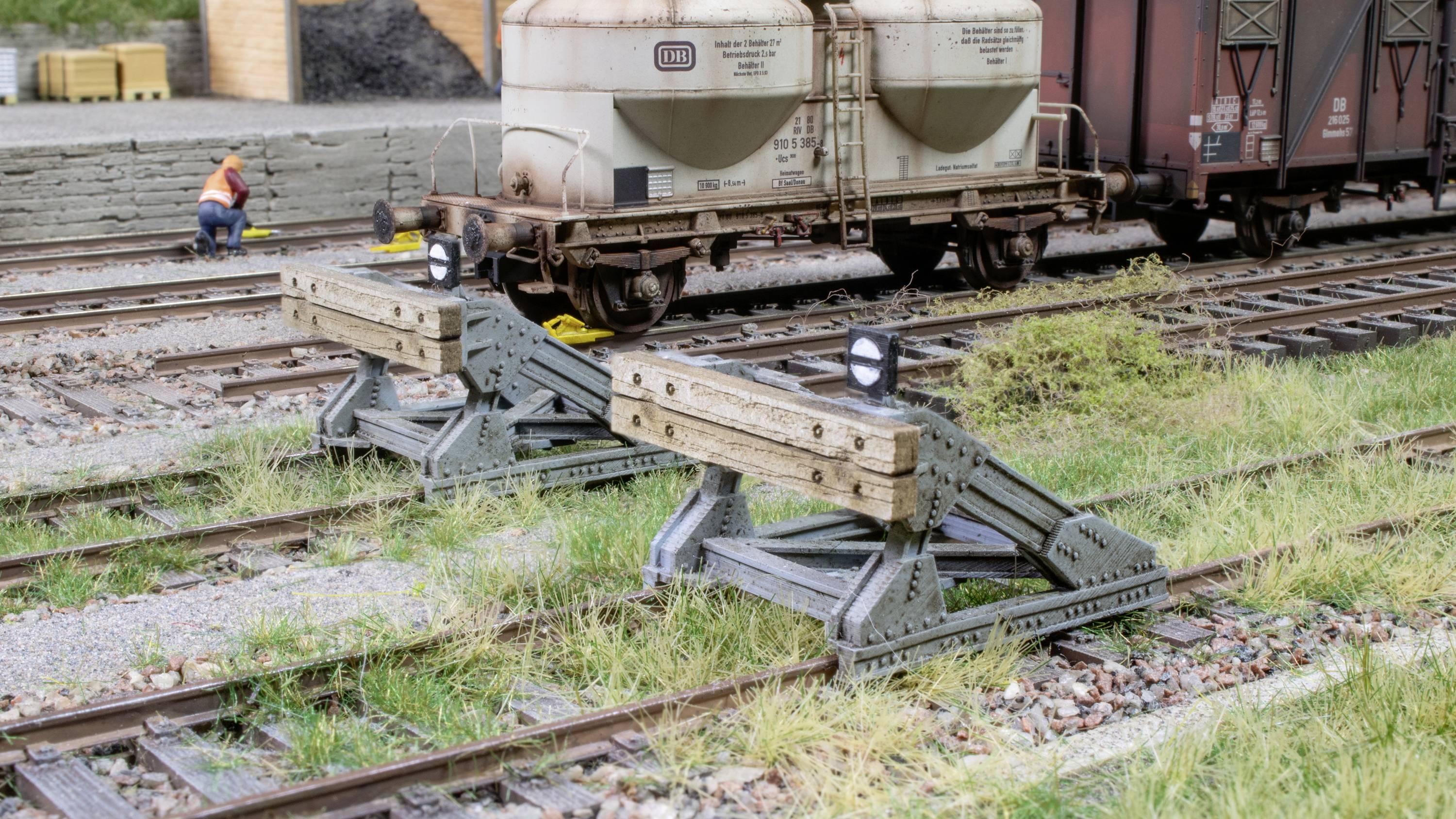 Zwei Prellböcke stoppen Schienen auf einem Bahngelände, dahinter stehende Güterwaggons. Ein Arbeiter in orangefarbener Weste im Hintergrund.