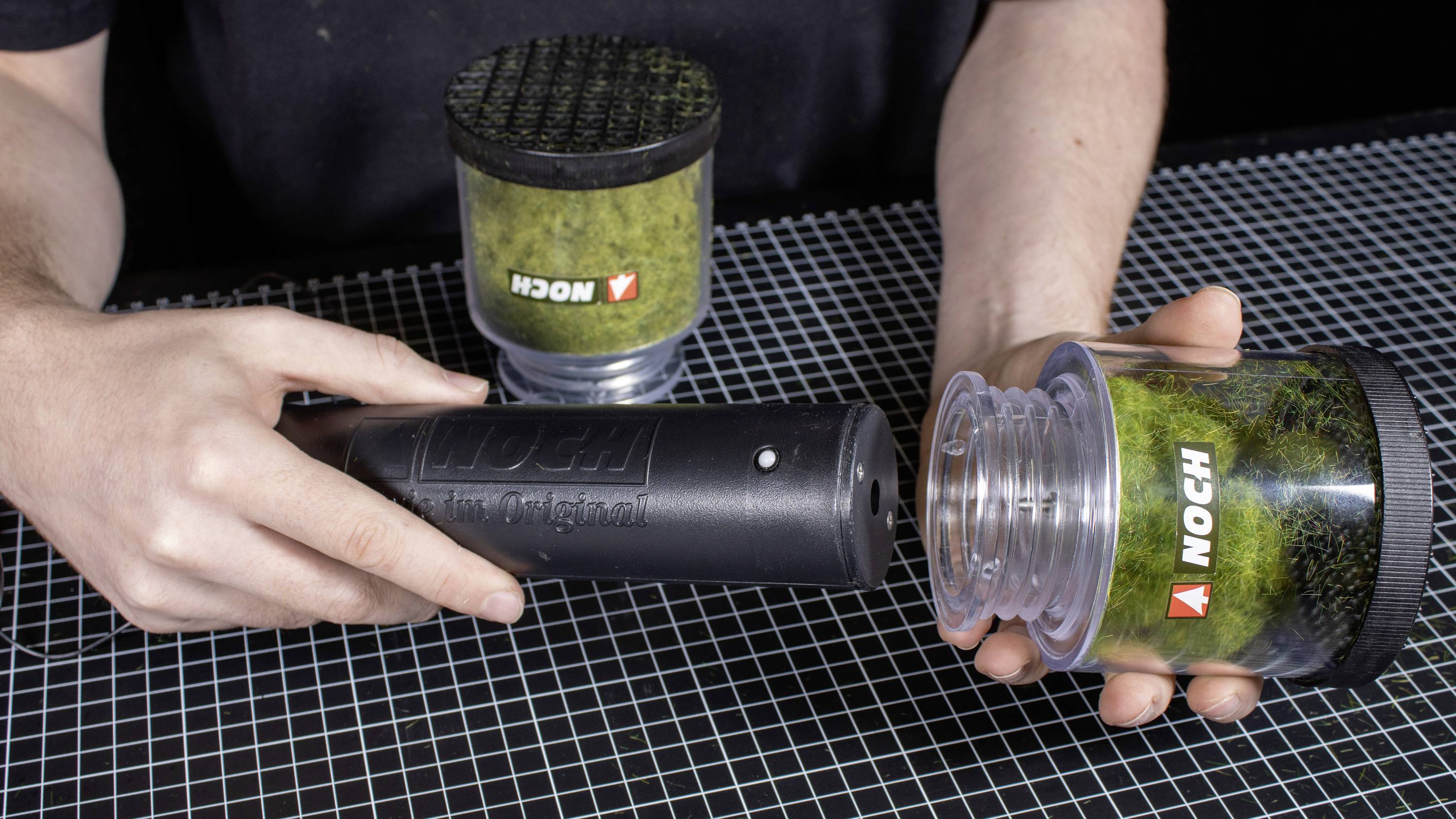 Eine Person zeigt, wie man Moosgrasstreu aus einem Behälter mit einem elektrostatischen Applikator aufträgt, um Modelllandschaften zu gestalten.