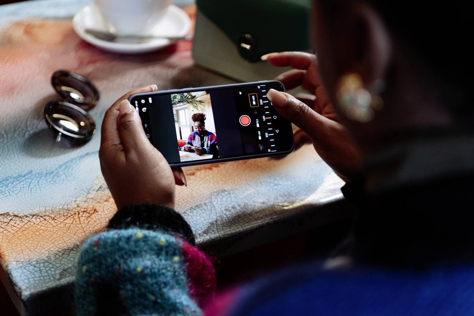 Person hält Smartphone und macht ein Foto in einem Café. Sonnenbrille und Kaffeetasse auf dem Tisch.