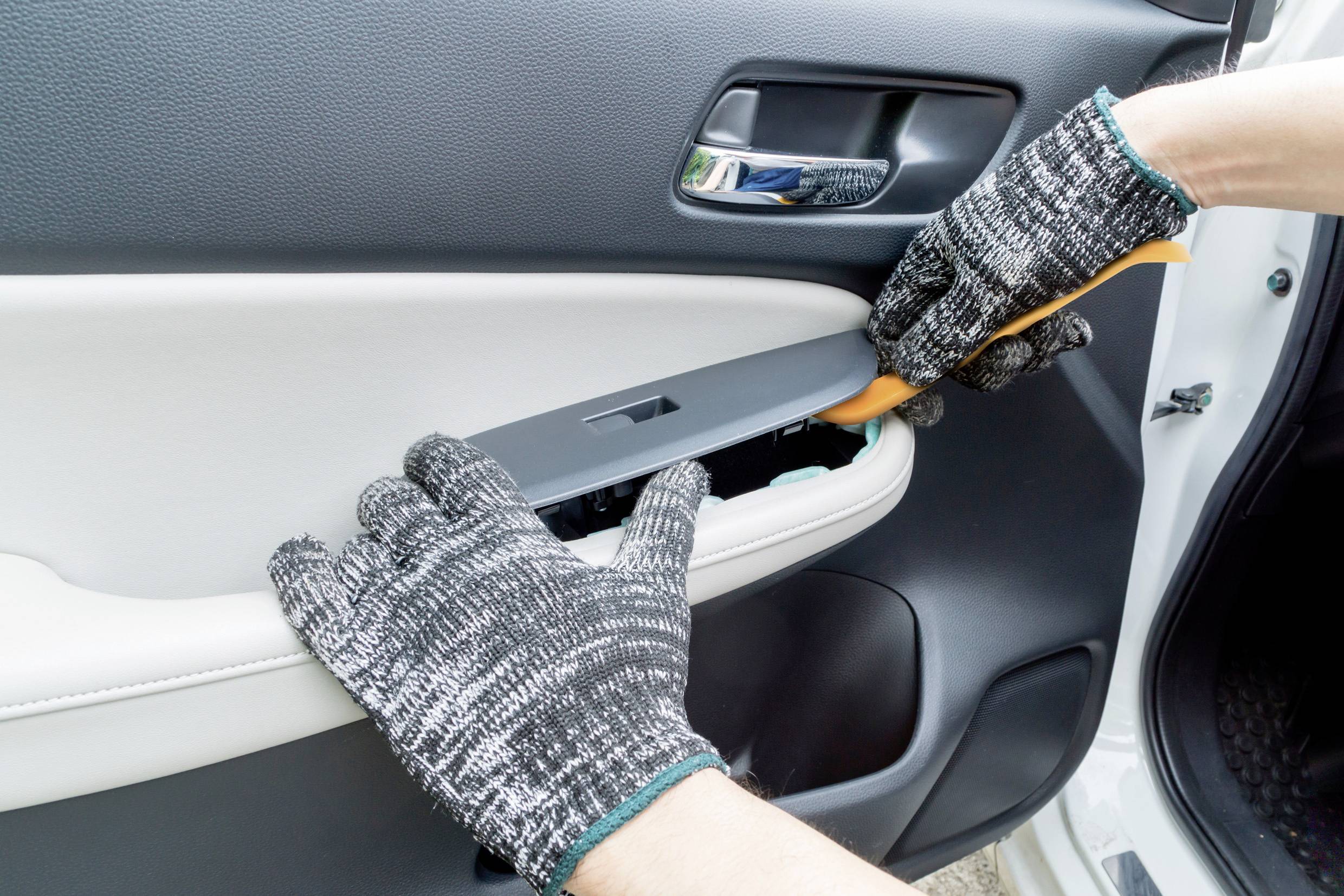 Zwei mit Handschuhen bekleidete Hände entfernen die Verkleidung eines Autotürgriffs mit einem Werkzeug.