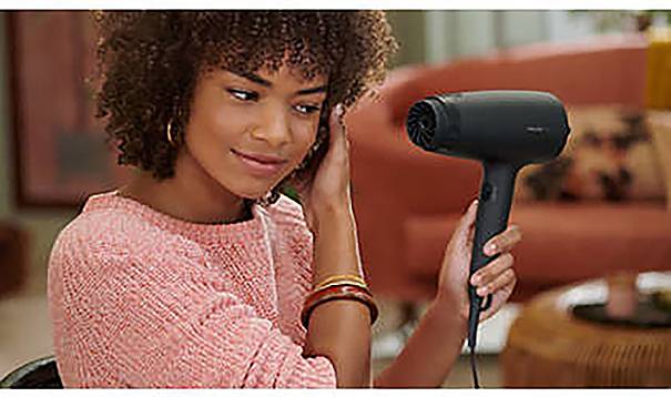 Eine Person mit lockigem Haar benutzt einen Haartrockner, trägt einen rosa Pullover und sitzt in einem Wohnraum.