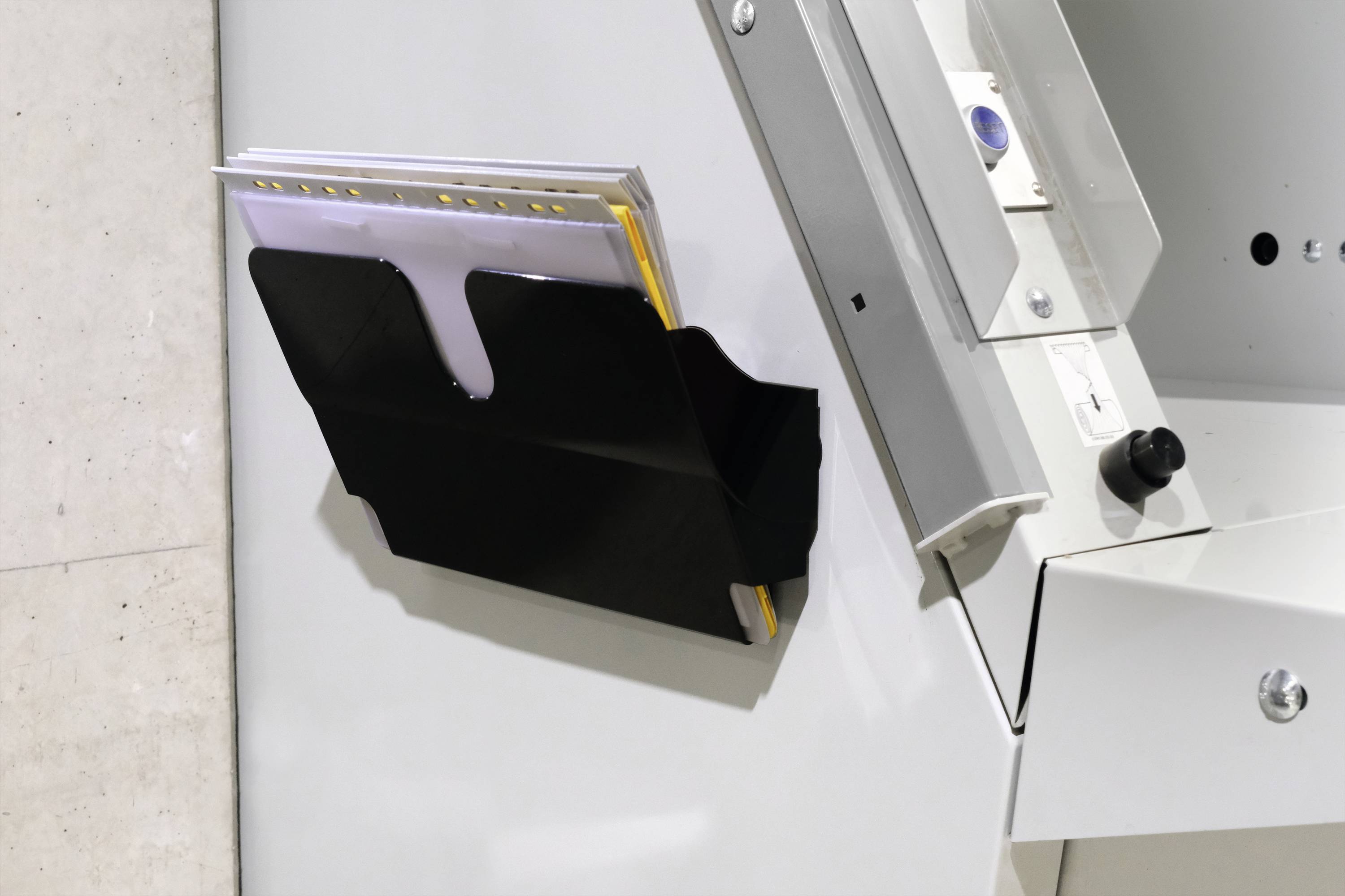 Schwarze Halterung mit Ordnern an einer silbernen Wand angebracht, nahe einer Maschine.