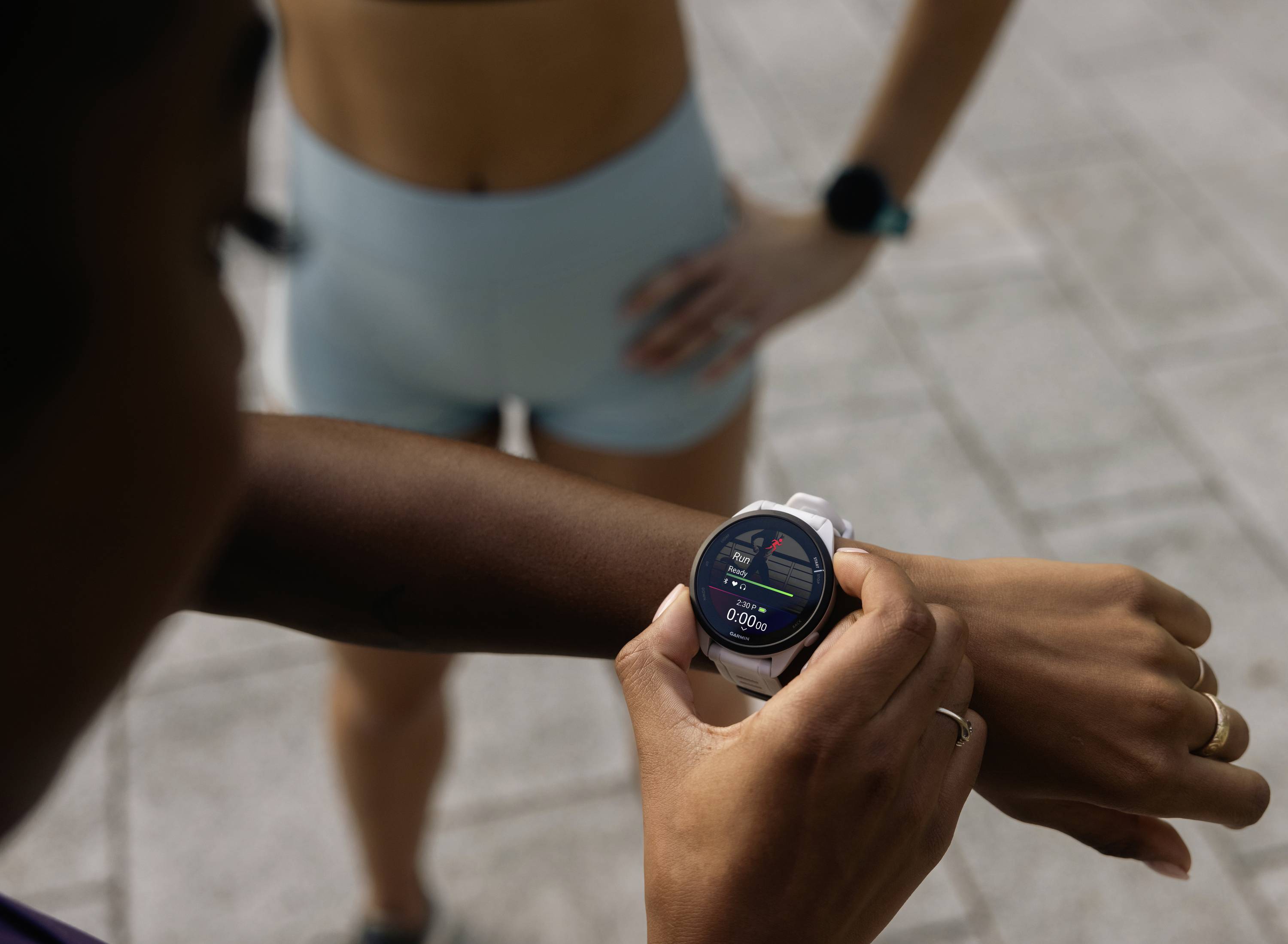 Person mit Smartwatch überwacht Fitnessdaten; zweite Person im Hintergrund in Sportkleidung. Hauptfokus auf Technik im Sport.