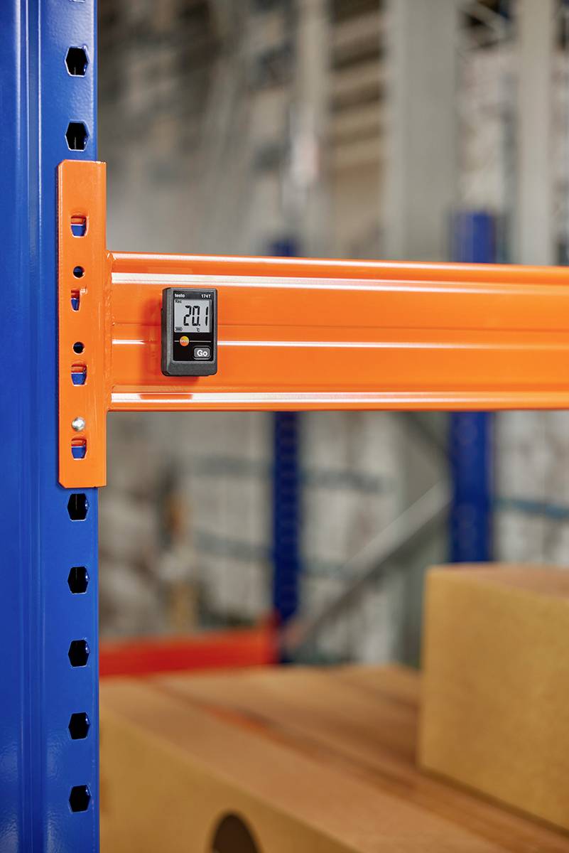 Ein digitales Thermometer zeigt 20 Grad Celsius an, befestigt an einem orangefarbenen Regal in einem Lager. Im Hintergrund sind Kartons sichtbar.