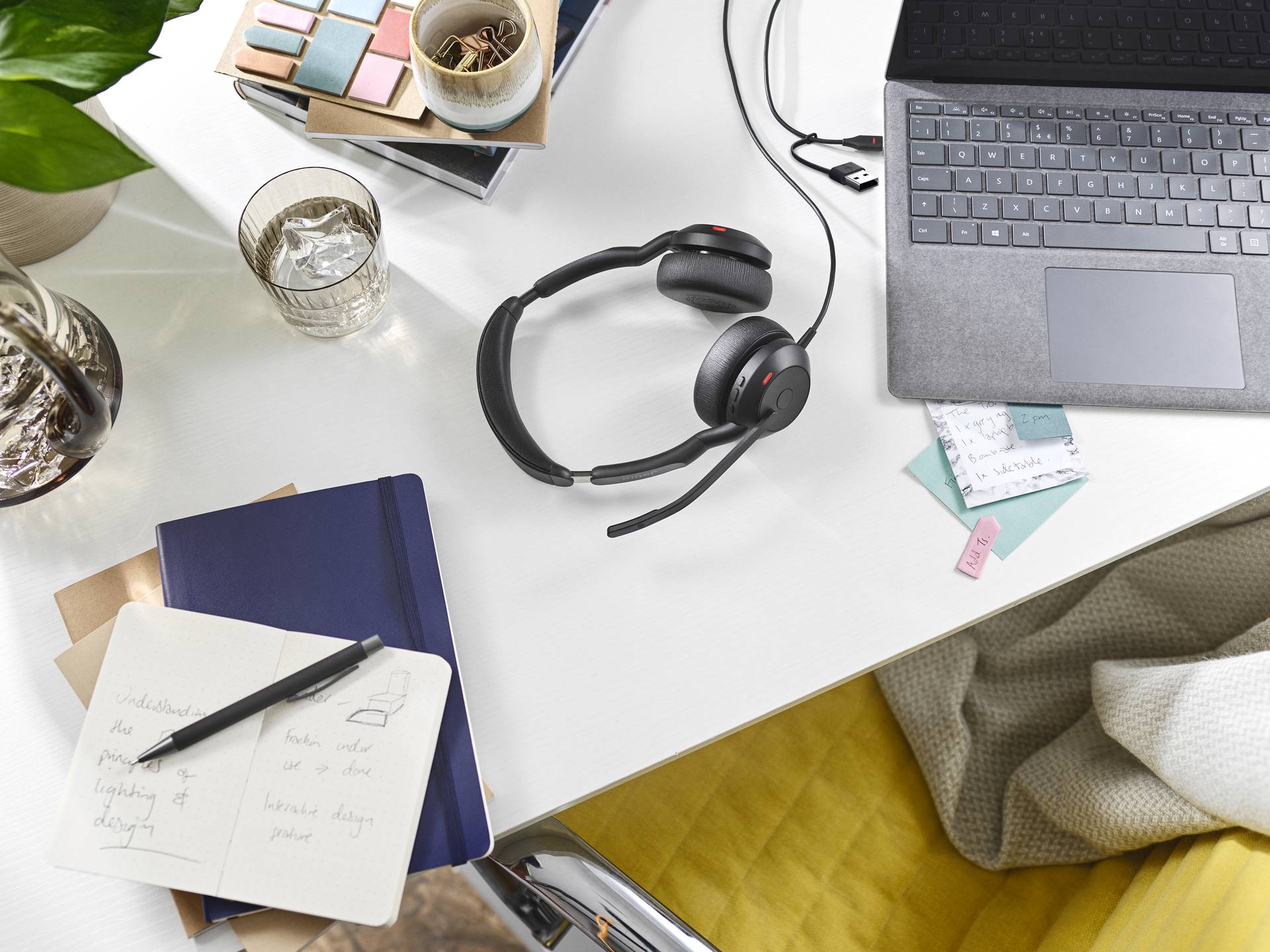 Ein Schreibtisch mit Laptop, Headset, Notizbuch und Glas Wasser; ein gemütlicher Arbeitsplatz mit Schreibmaterialien und Pflanzen.