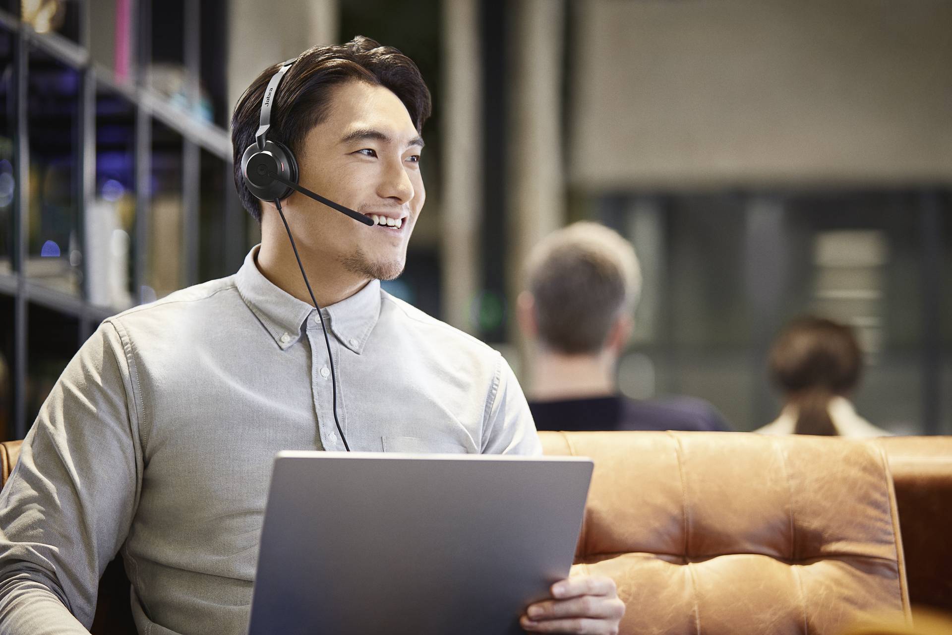 Ein Mann mit Headset sitzt auf einem Sofa, hält einen Laptop und lächelt, während er in einem Büro arbeitet. Im Hintergrund unscharfe Personen.