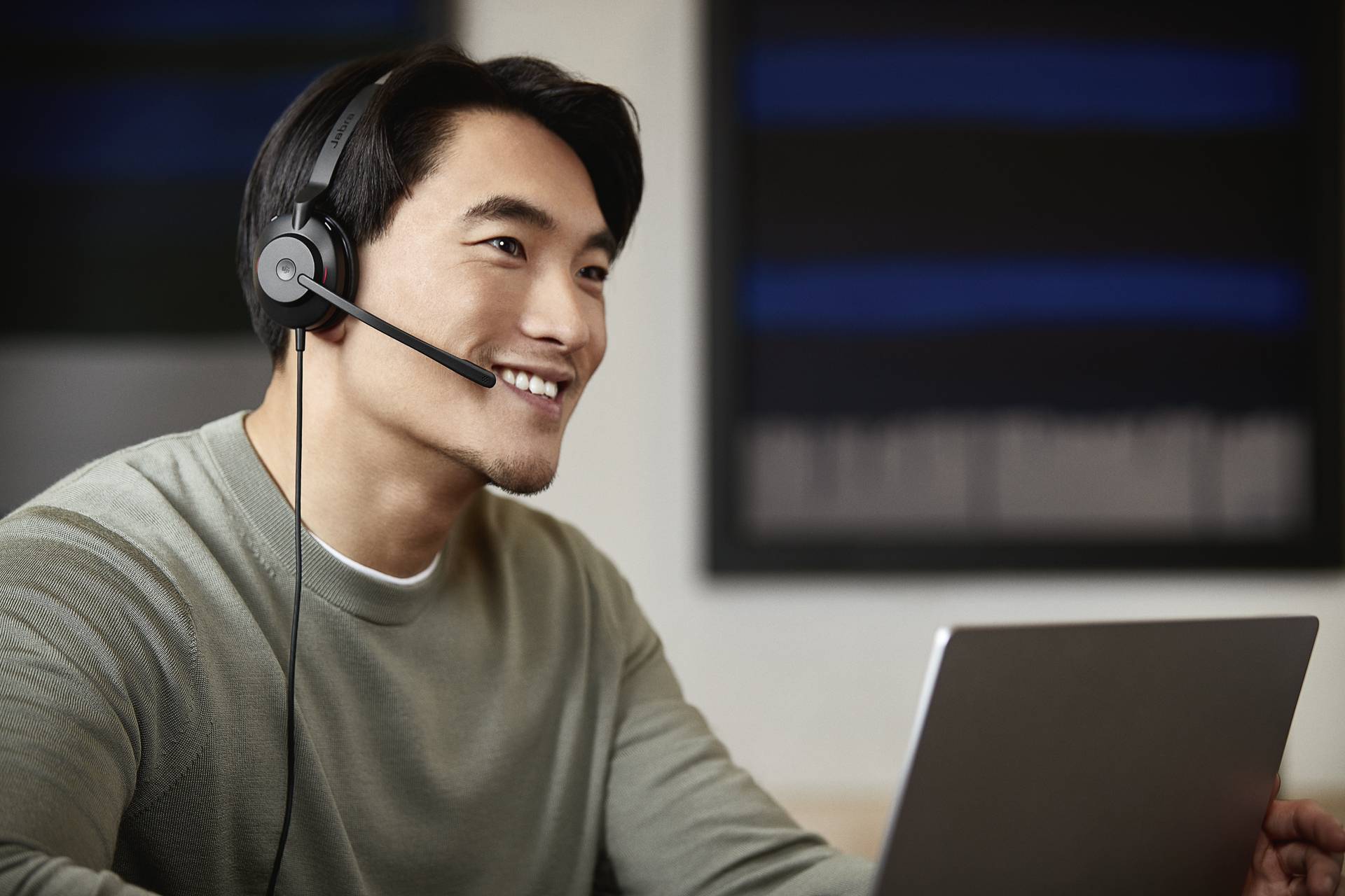 Mann mit Headset und Laptop lächelt bei der Arbeit. Hintergrund unscharf, zeigt Büro- oder Arbeitsumgebung.