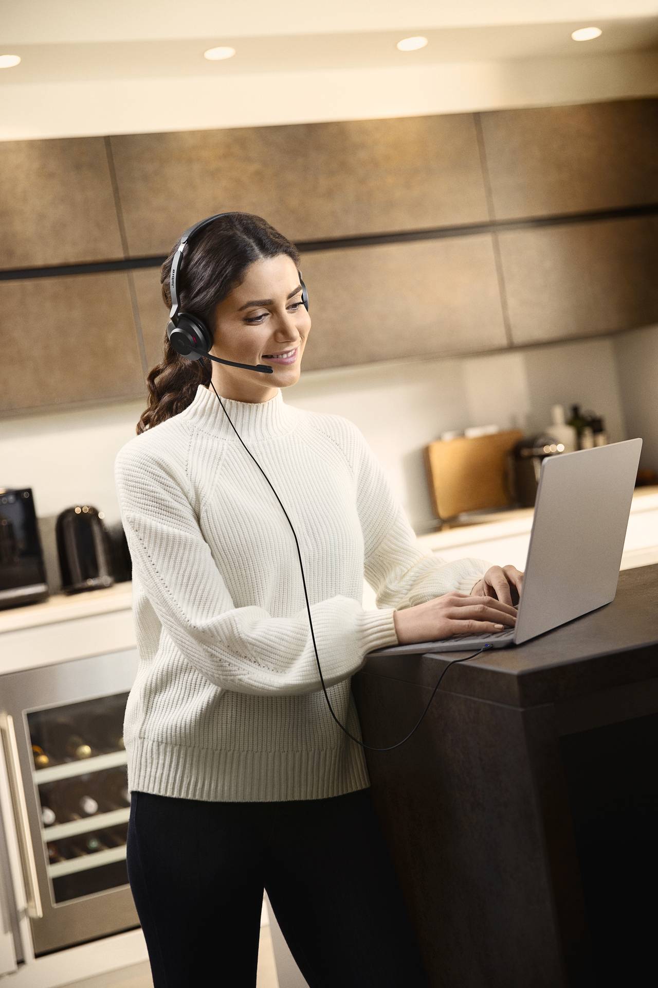 Eine Frau mit Headset steht in einer modernen Küche und tippt auf einem Laptop. Sie wirkt konzentriert und trägt einen weißen Pullover.