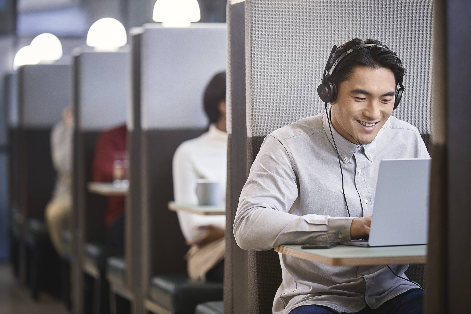 Ein Mann mit Kopfhörern, lächelnd, sitzt an einem Tisch in einem Café und arbeitet an einem Laptop. Im Hintergrund sind weitere Personen zu sehen.