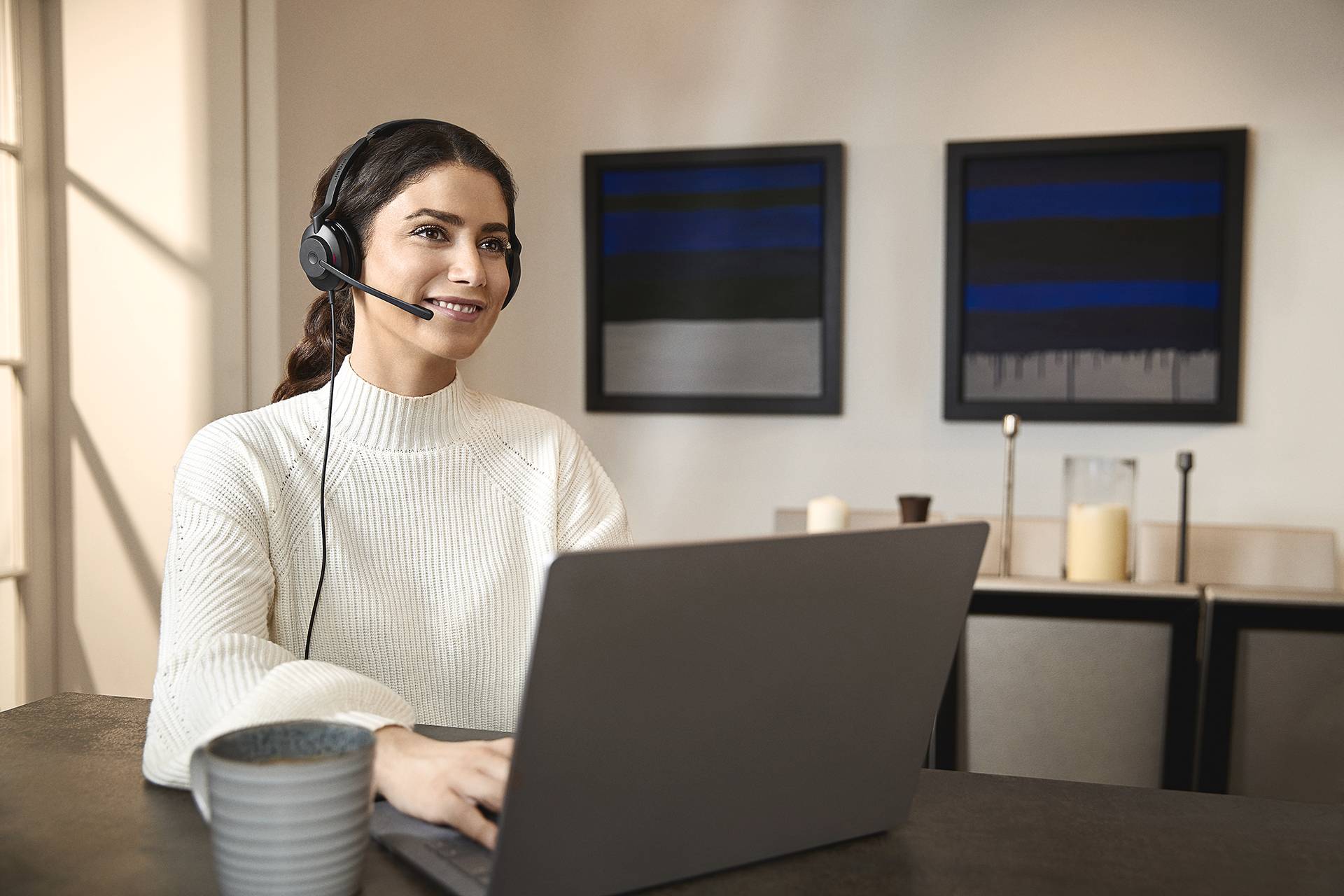 Eine Frau sitzt mit einem Headset lächelnd vor einem Laptop in einem modernen Raum. Im Hintergrund sind abstrakte Gemälde sichtbar.