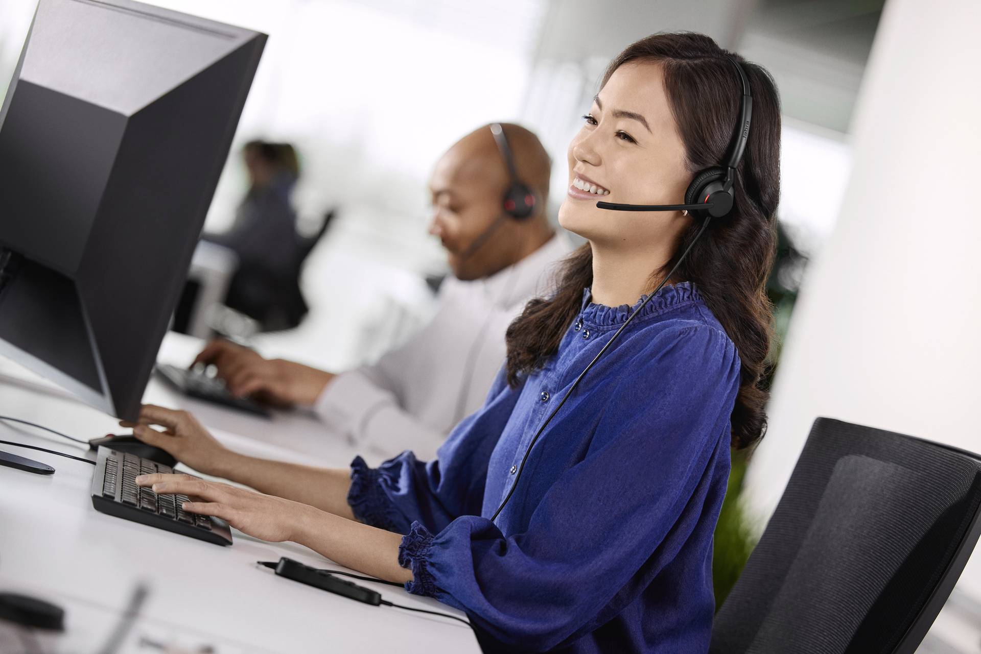 Eine Frau und ein Mann arbeiten an Computern in einem Büro. Beide tragen Headsets und lächeln, während sie auf Bildschirme schauen.