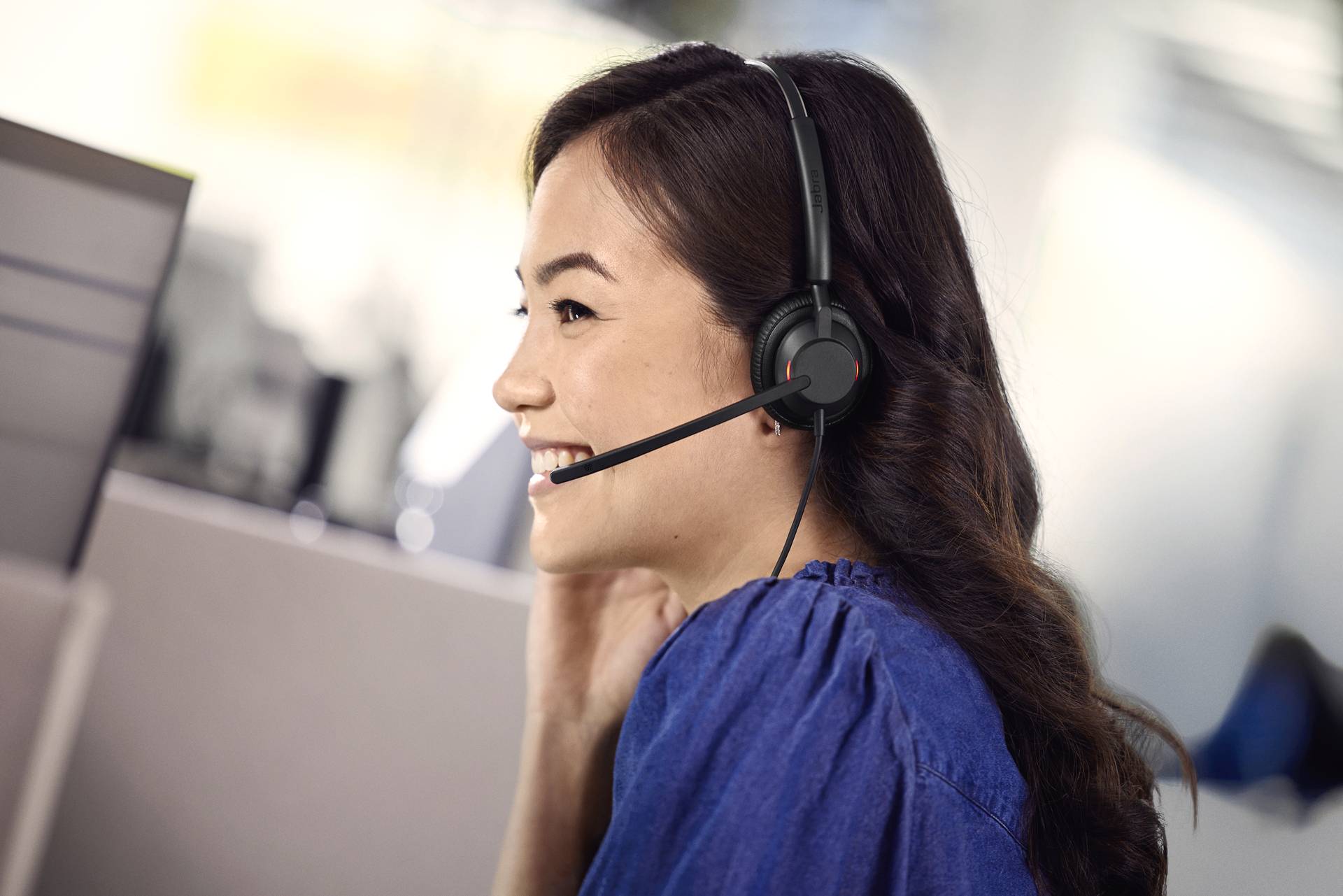 Eine Frau mit einem Headset lächelt und spricht. Sie sitzt in einem modernen Büro, möglicherweise in einem Kundenservice- oder Support-Center.