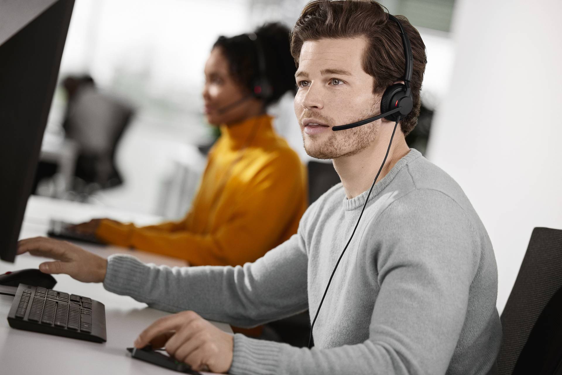 Ein Mann mit Headset sitzt an einem Computer und spricht, wahrscheinlich in einem Callcenter. Im Hintergrund ist eine weitere Person zu sehen.
