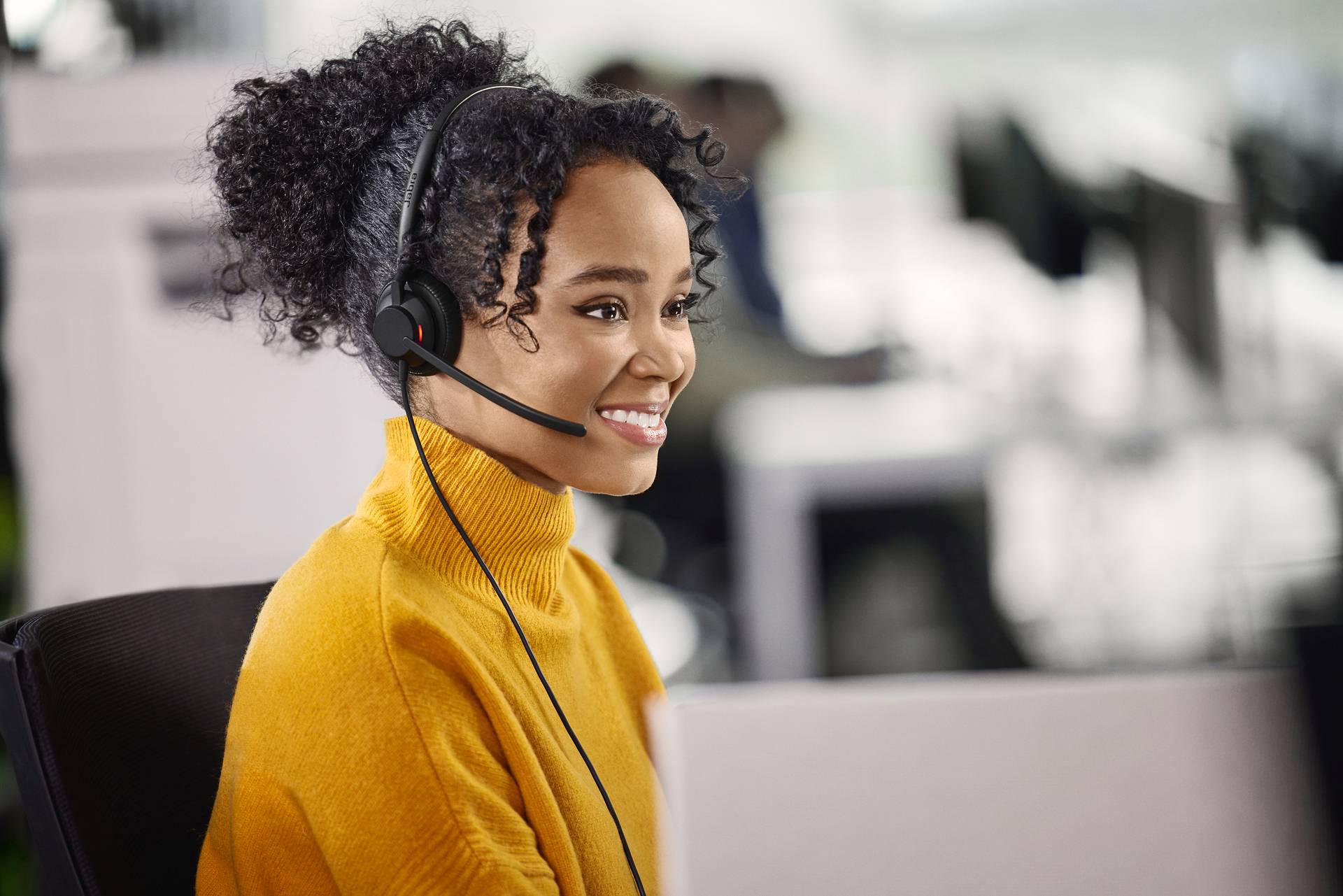 Eine Person mit einem Headset sitzt an einem Schreibtisch und lächelt. Der Hintergrund zeigt ein geschäftiges Büro.