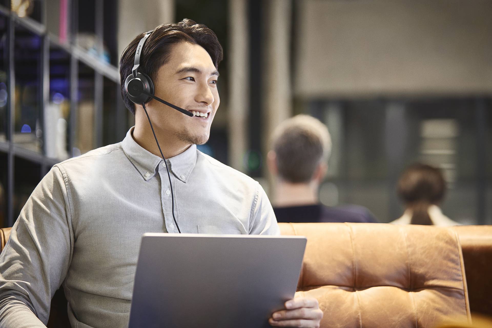 Ein Mann mit Headset sitzt auf einem Ledersofa und hält einen Laptop. Im Hintergrund sind verschwommene Personen zu sehen.